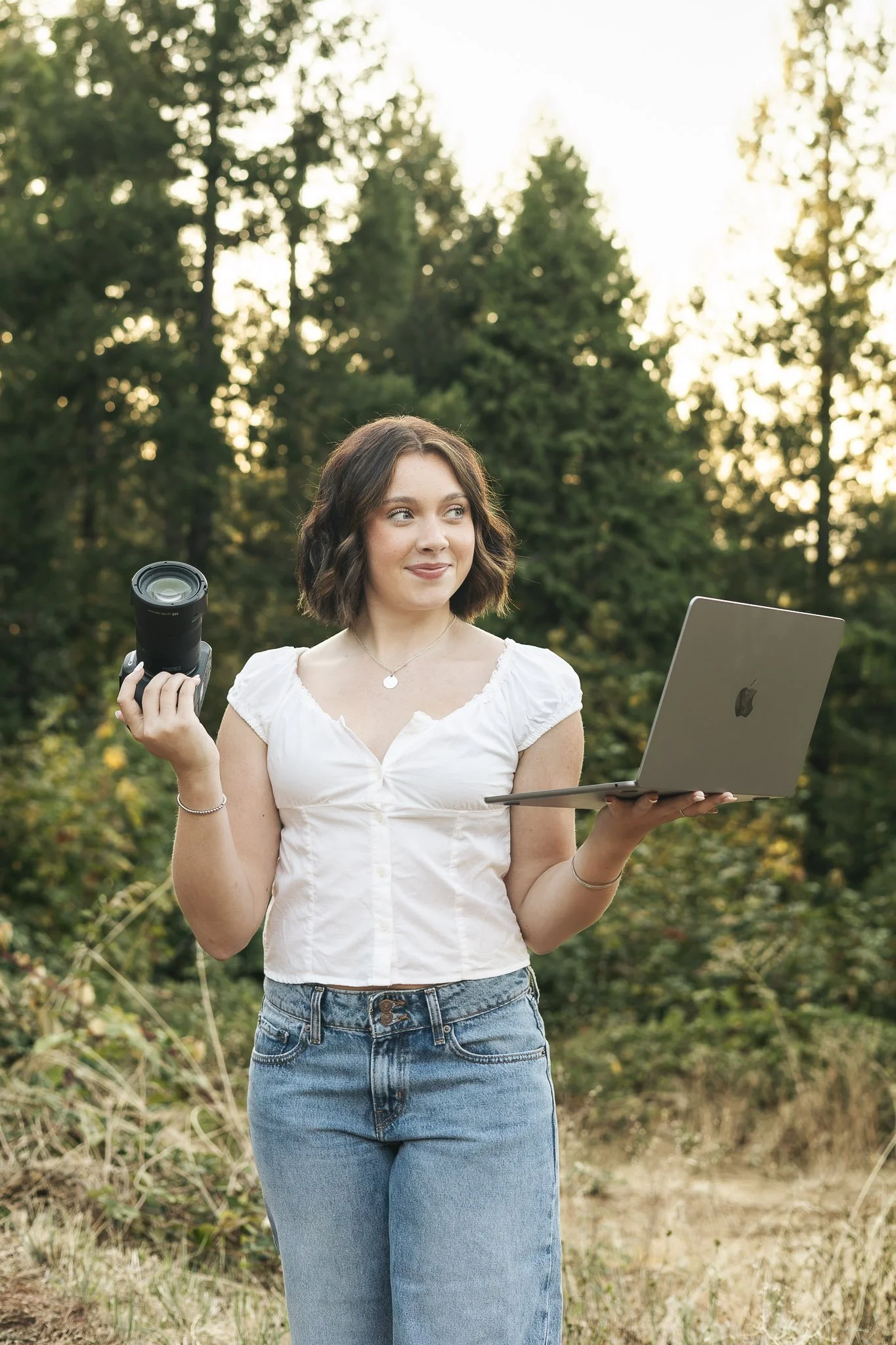A young woman with shoulder-length brown hair holding a camera in one hand and a laptop in the other, standing outdoors in a forested area with sunlight filtering through trees.