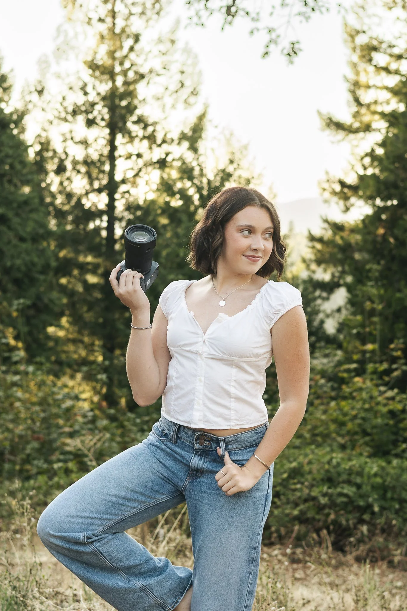 A young woman with short brown hair standing outside in a natural setting, holding a camera, wearing a white blouse and blue jeans, with trees and sunlight in the background.