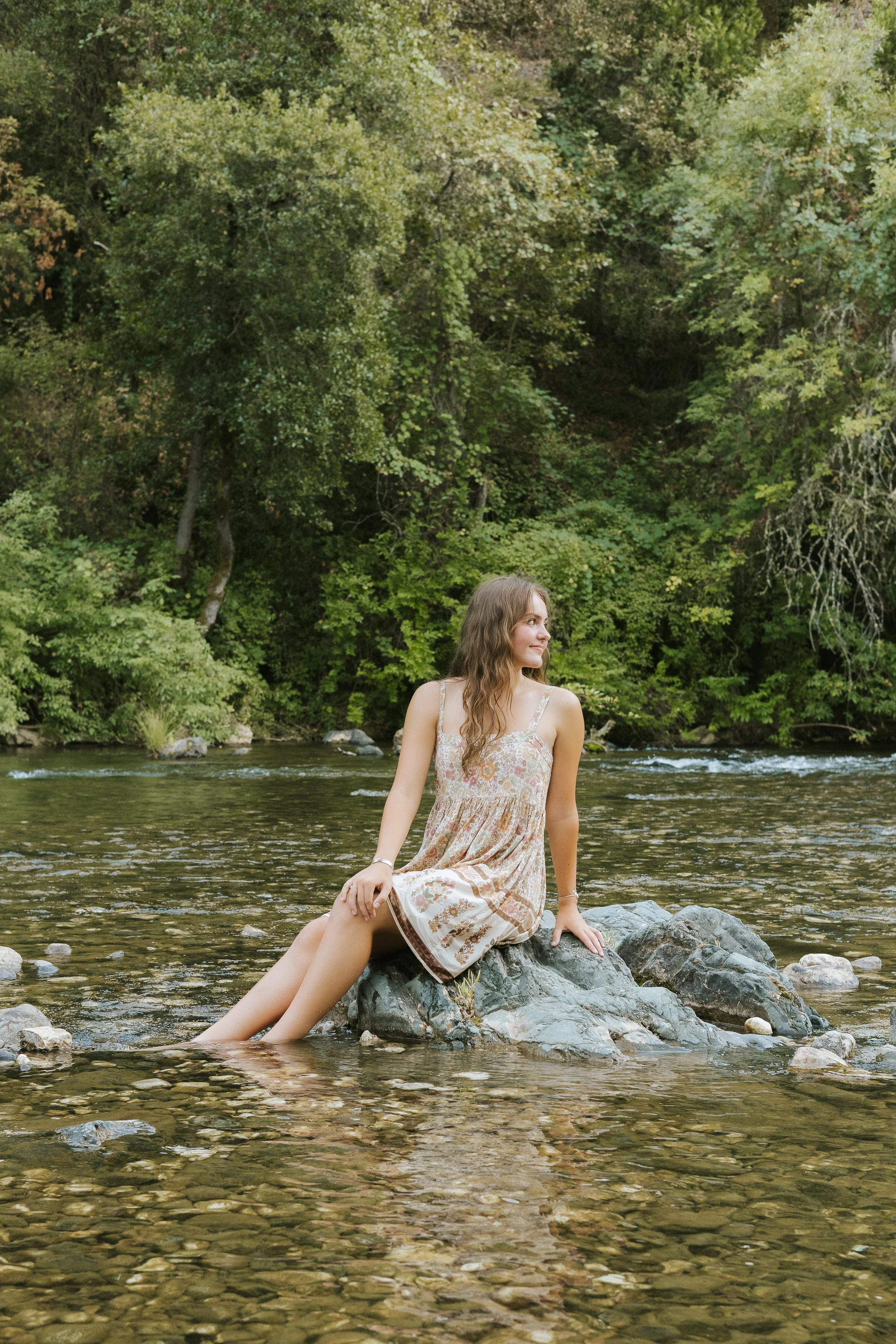 A young woman with long brown hair sitting on a rock in the middle of a river, surrounded by green trees.