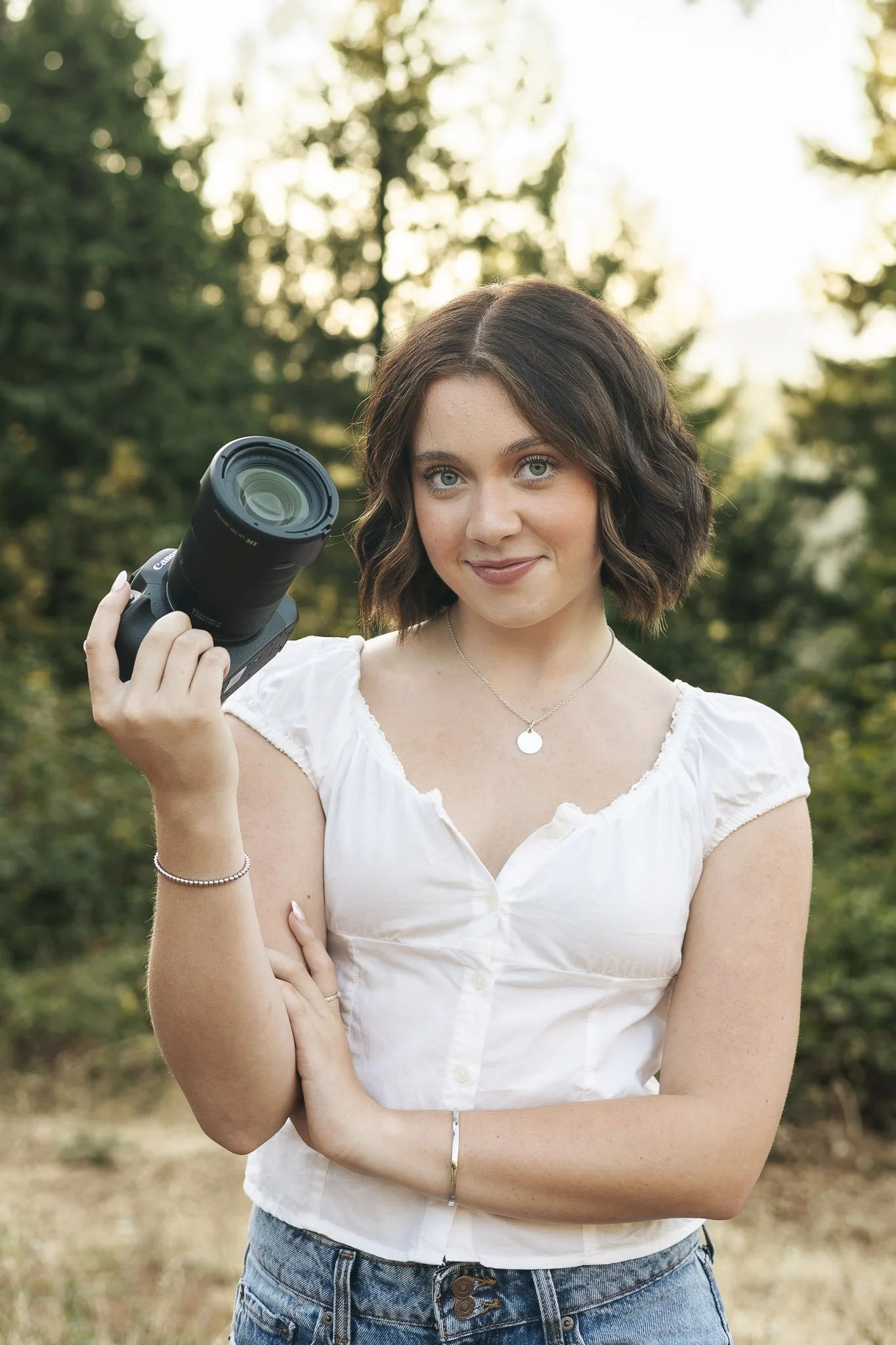 A woman with short curly brown hair and blue eyes holding a camera outdoors with a blurred background of trees and sunlight.