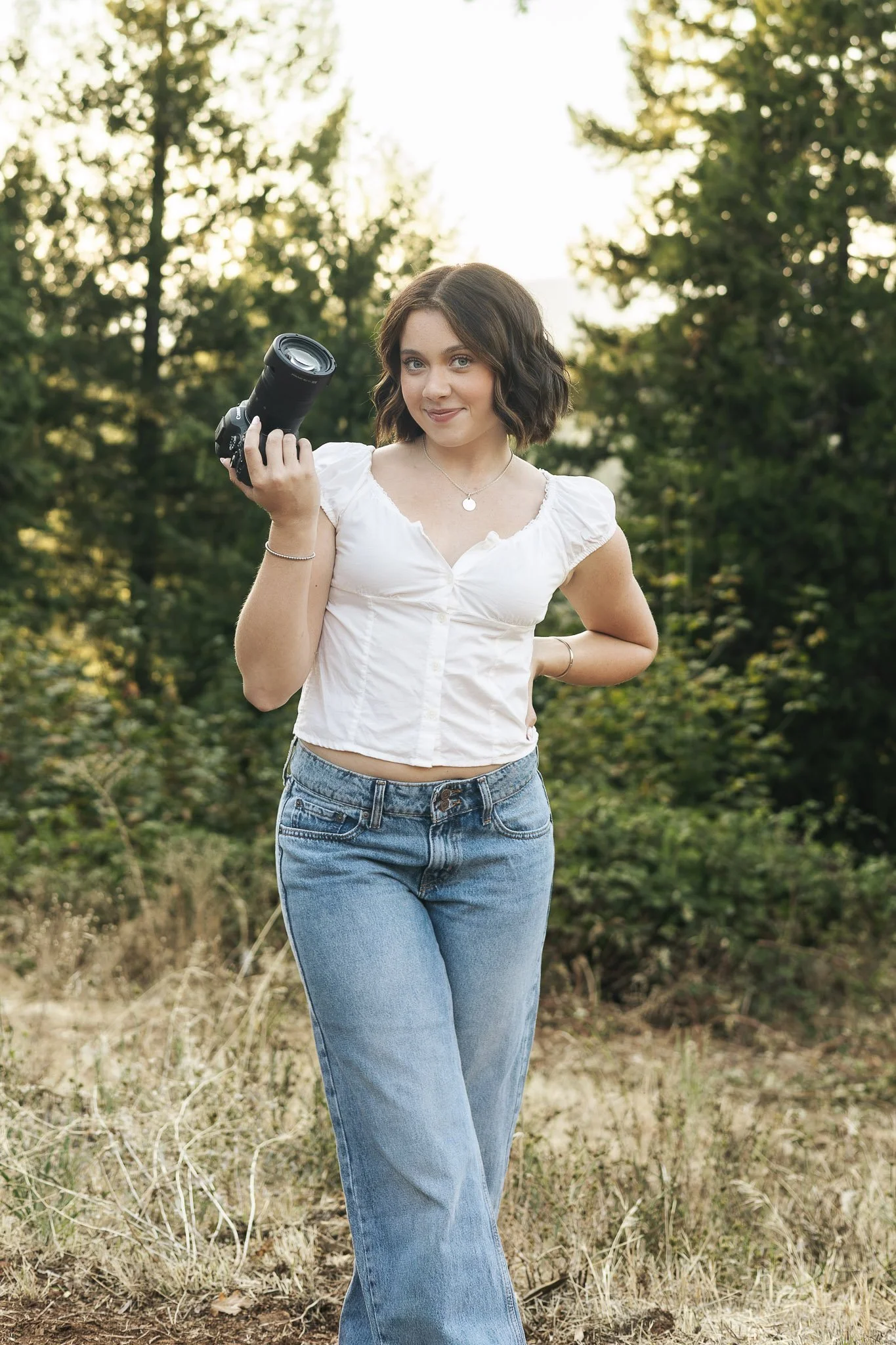 A young woman with short wavy brown hair and blue eyes, wearing a white blouse and blue jeans, stands outdoors holding a camera against a background of green trees and sunlight.