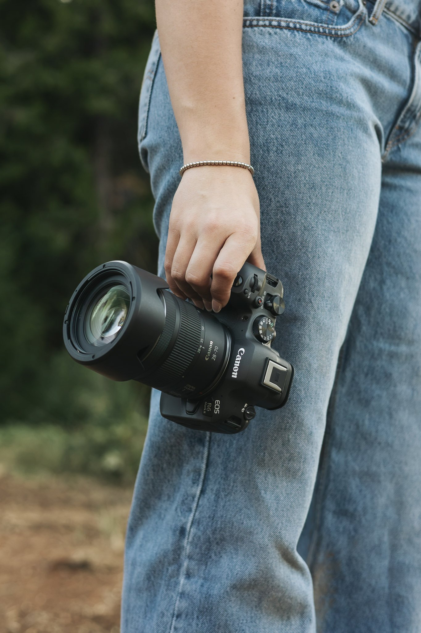 Person in blue jeans holding a camera outdoors, with a blurred green background.