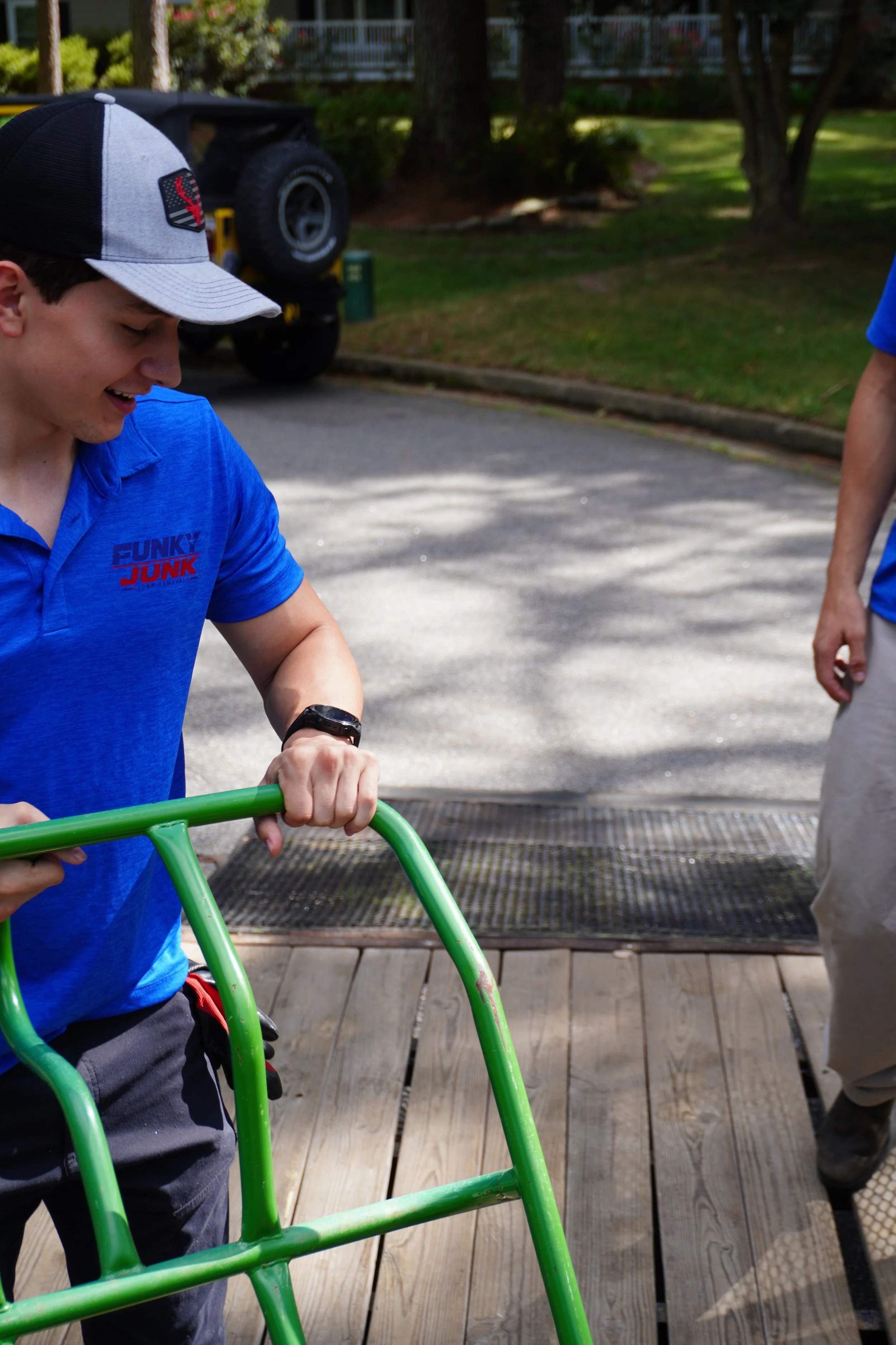 A young man wearing a blue shirt with the logo 'FUNKY JUNK' on it, a gray and black baseball cap, and a watch, is holding a green object, possibly a part of a bike or outdoor equipment, on a wooden deck. There is a tree, grass, a trailer with a spare tire, and a house in the background.