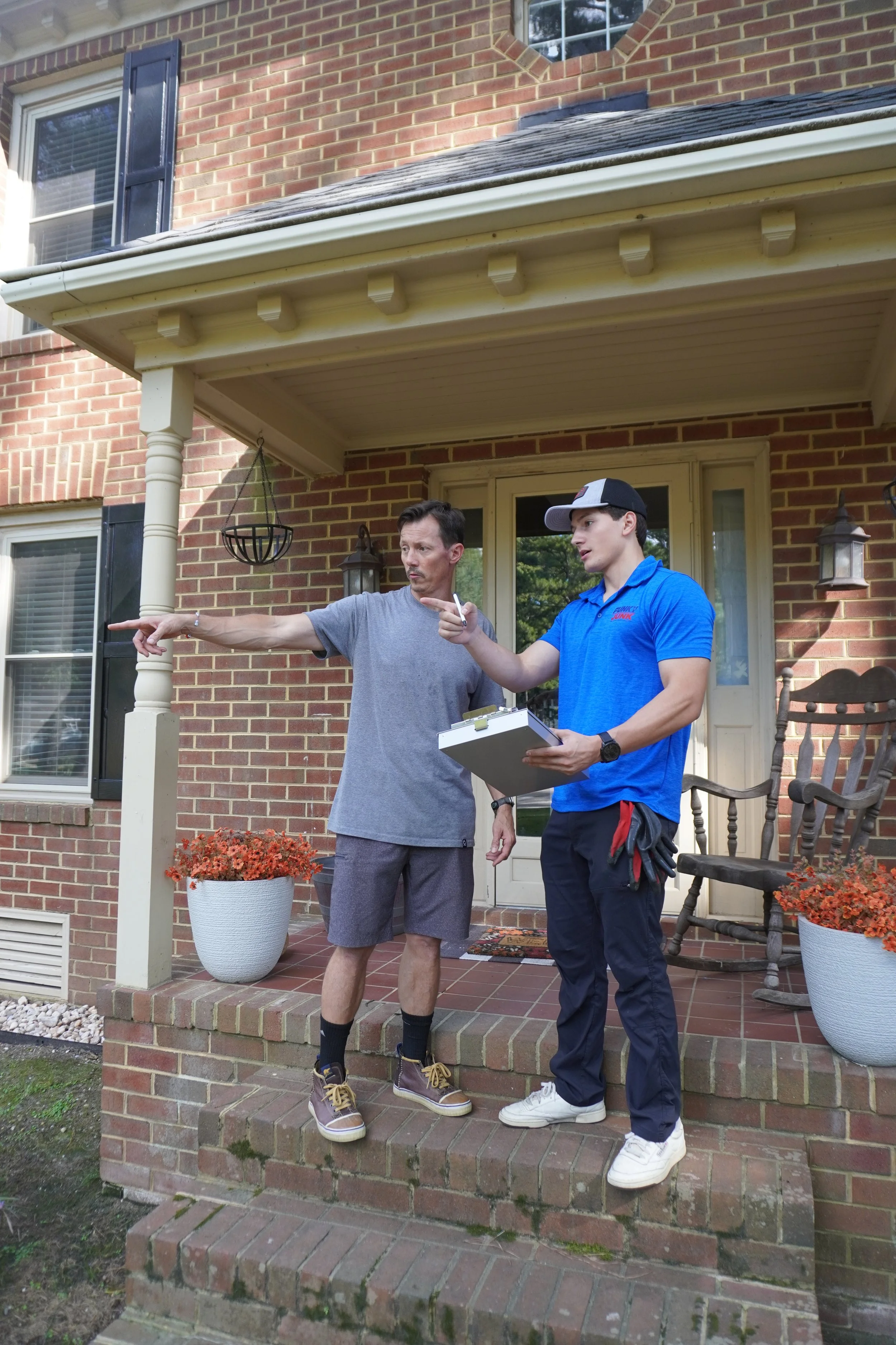 Two men standing on the front porch of a brick house, engaged in a conversation. One man points to something outside the frame. The other holds a clipboard and wears a hat. The porch has two large potted plants, a hanging basket, and an outdoor light fixture.