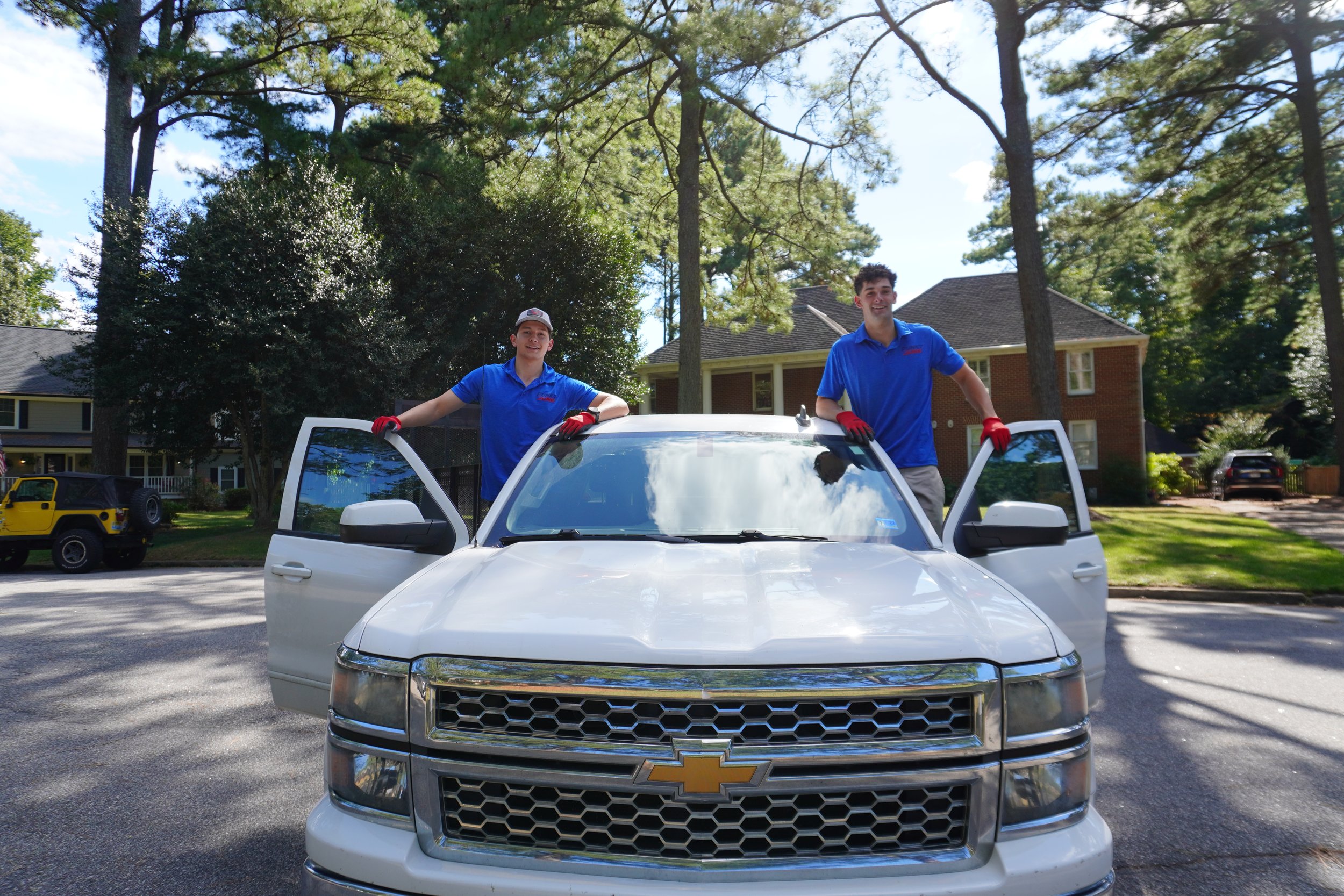 Two young men in blue shirts and red gloves standing in an open white Chevrolet SUV with the driver and passenger doors open, outdoors on a suburban street with trees, houses, and a yellow toy vehicle in the background.