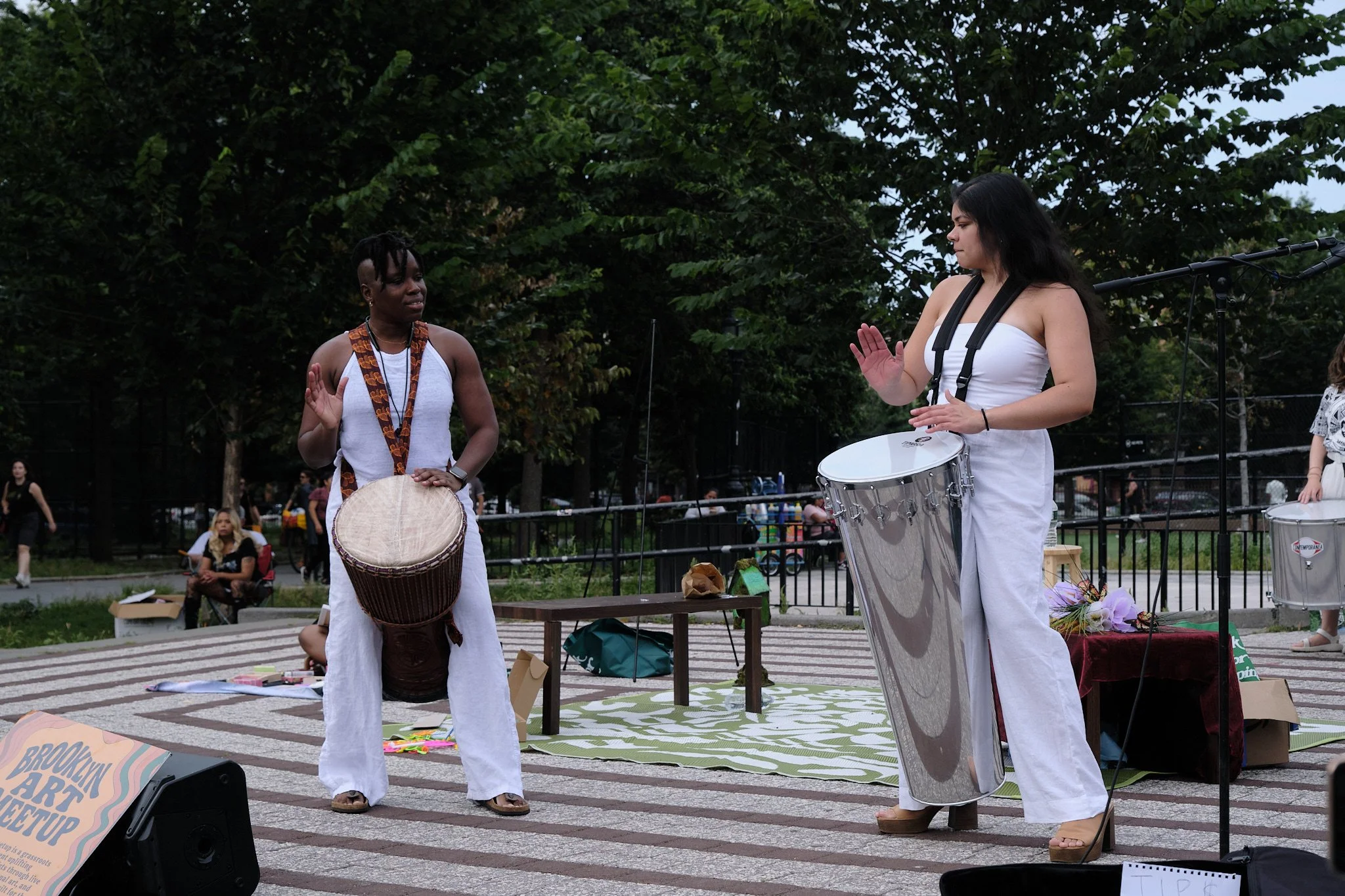 Two women in white clothing and sandals singing or performing with drums in a park, with people sitting on benches and watching in the background.