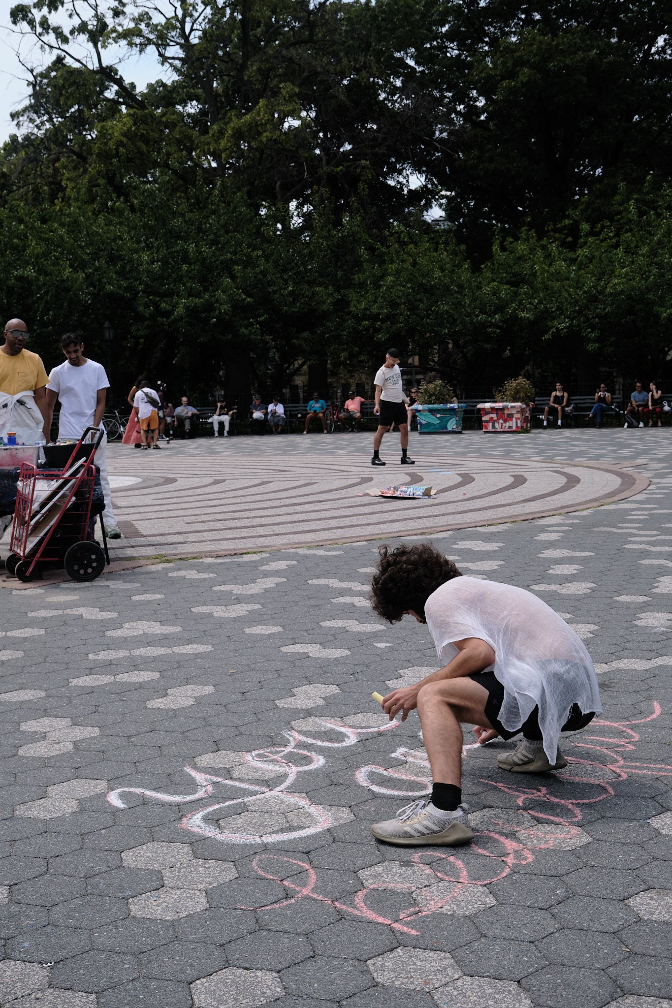 A man crouches on the ground drawing with chalk on a paved area in a park, surrounded by people sitting on benches and trees.
