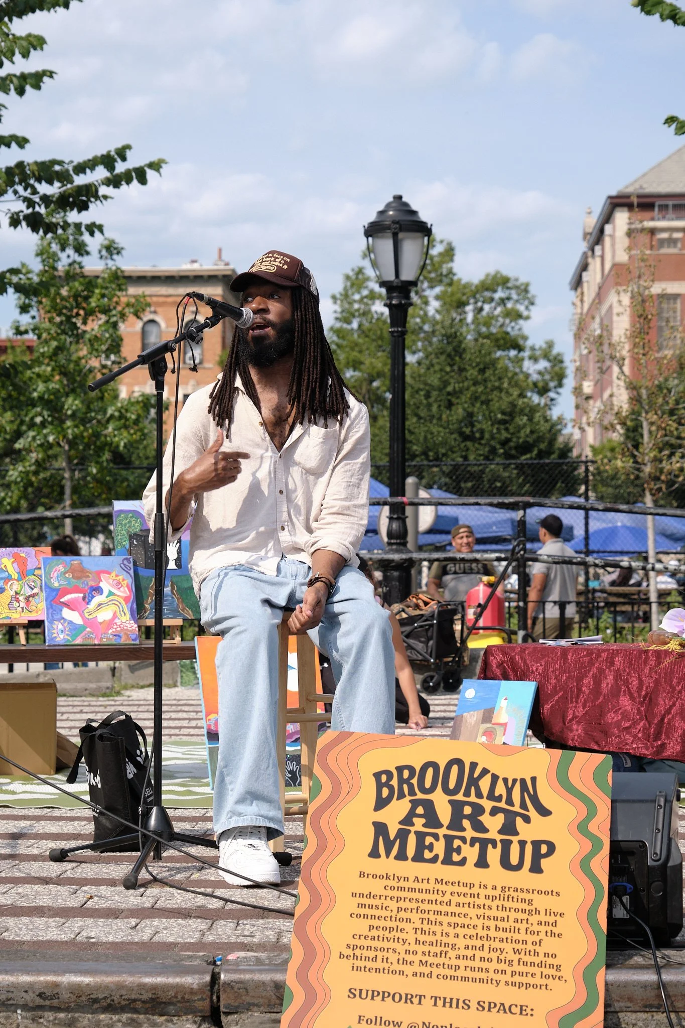 A man with long dreadlocks and a cap is speaking into a microphone at an outdoor art meetup event in Brooklyn. There are paintings and art supplies displayed around him, and a sign explaining the event.