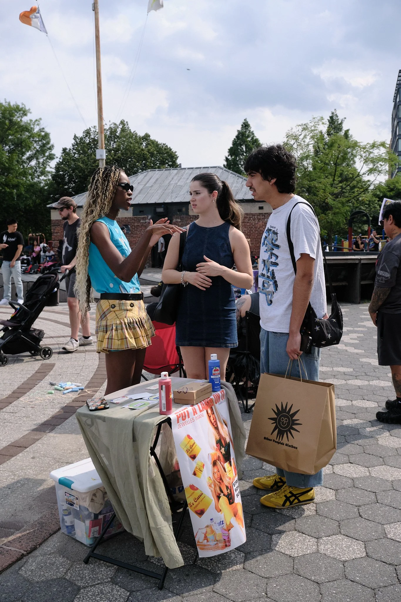 Two women and a man are having a conversation at an outdoor event. The women are standing behind a small table with health and beauty products, including bottles and a poster advertising beauty treatments. People and trees are visible in the backgrou
