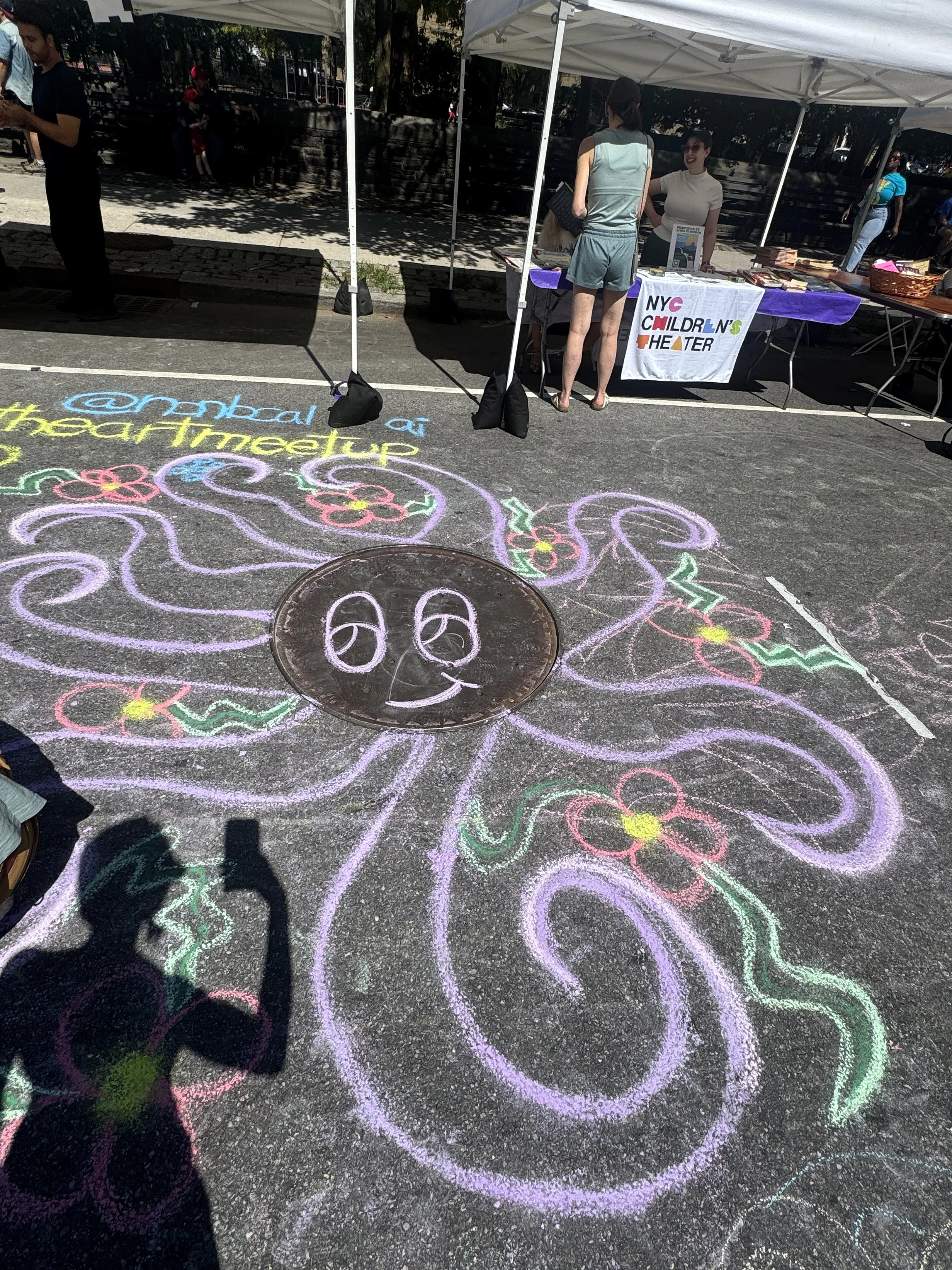 Colorful chalk drawings of flowers and swirls on a sidewalk at an outdoor event. Several people are visible under tents, with a sign reading 'NYC Children's Theater' on a table.