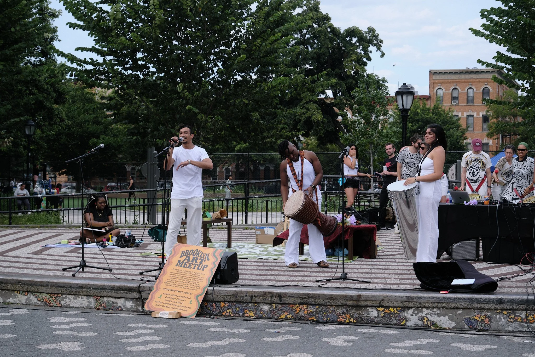 Group of performers and musicians on an outdoor stage in a park, with trees and brick buildings in the background, during a Brooklyn meet-up event.