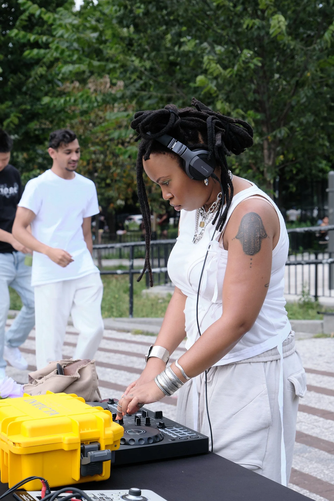A woman with dreadlocks wearing headphones, a white tank top, and jewelry is DJing outdoors at a park. Two men in white and black shirts are in the background, and there are trees and a fence.