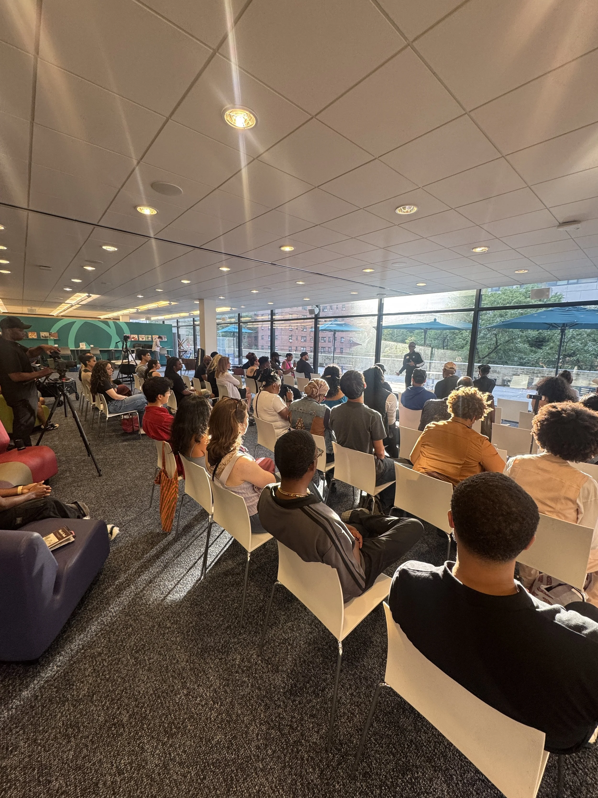A large group of people seated in a modern conference room with floor-to-ceiling windows, listening to a speaker at the front, with a cityscape visible outside.
