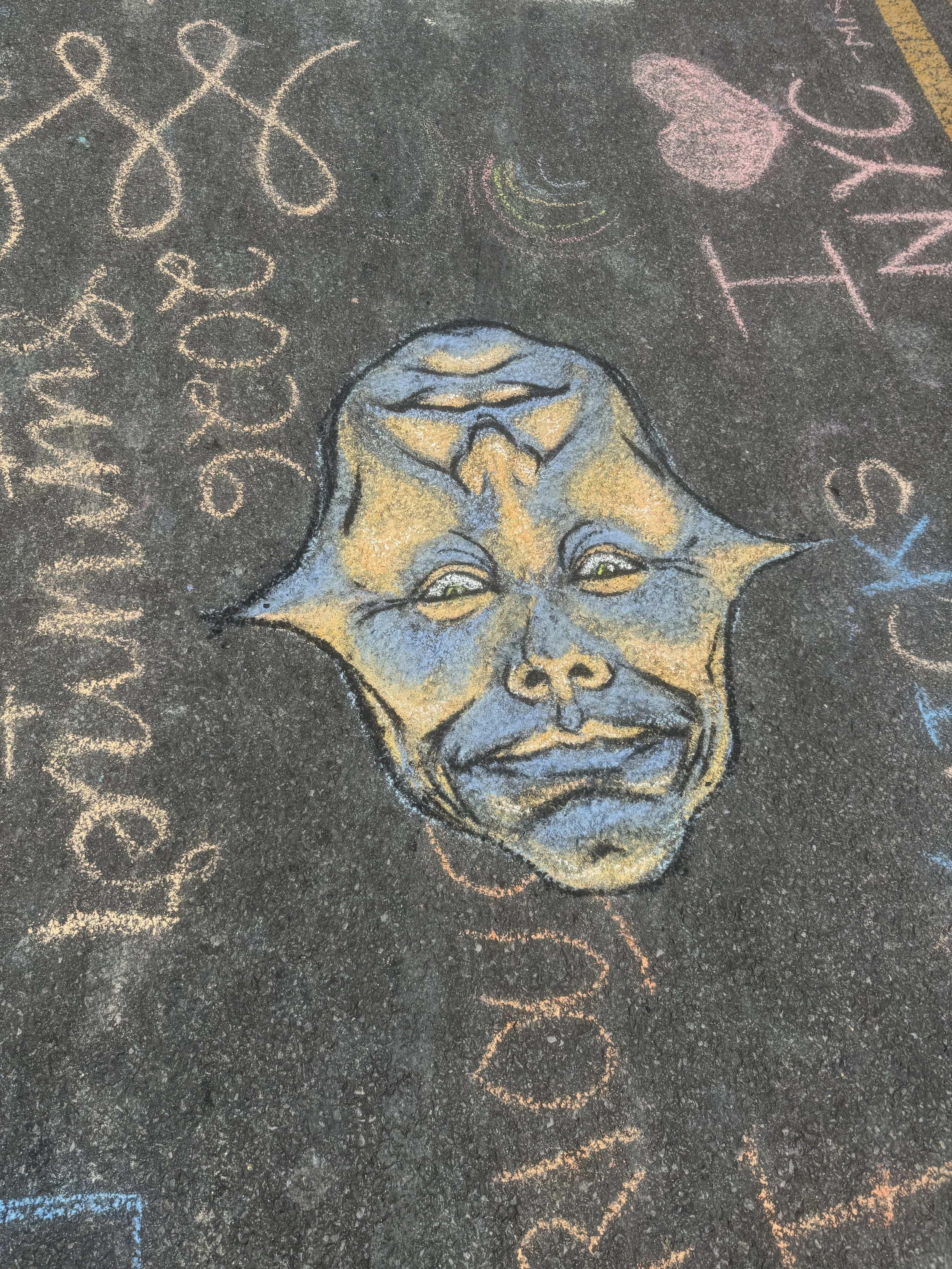 Chalk drawing on asphalt of a child's face with glasses and a scarecrow hat, along with colorful words and hearts.