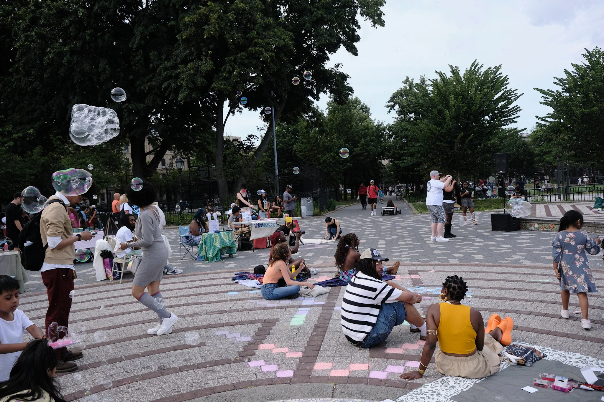 People gathered in a park for an outdoor event, some sitting on the ground, others walking and standing. Children and adults are present, with some playing with soap bubbles. There are trees and a fence in the background, and booths or tables set up 