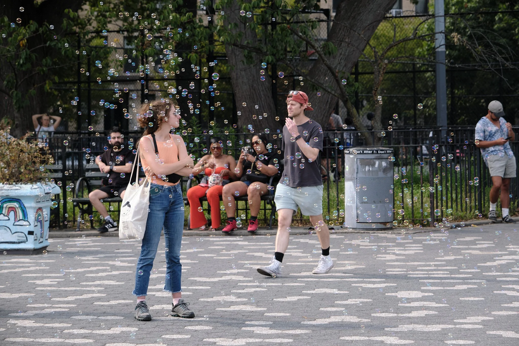 A scene in a park where two young people, a woman and a man, are walking through a bubble display. The woman is wearing glasses, a peach sleeveless top, and jeans, while the man is in a backwards baseball cap, gray t-shirt, and shorts. Several people