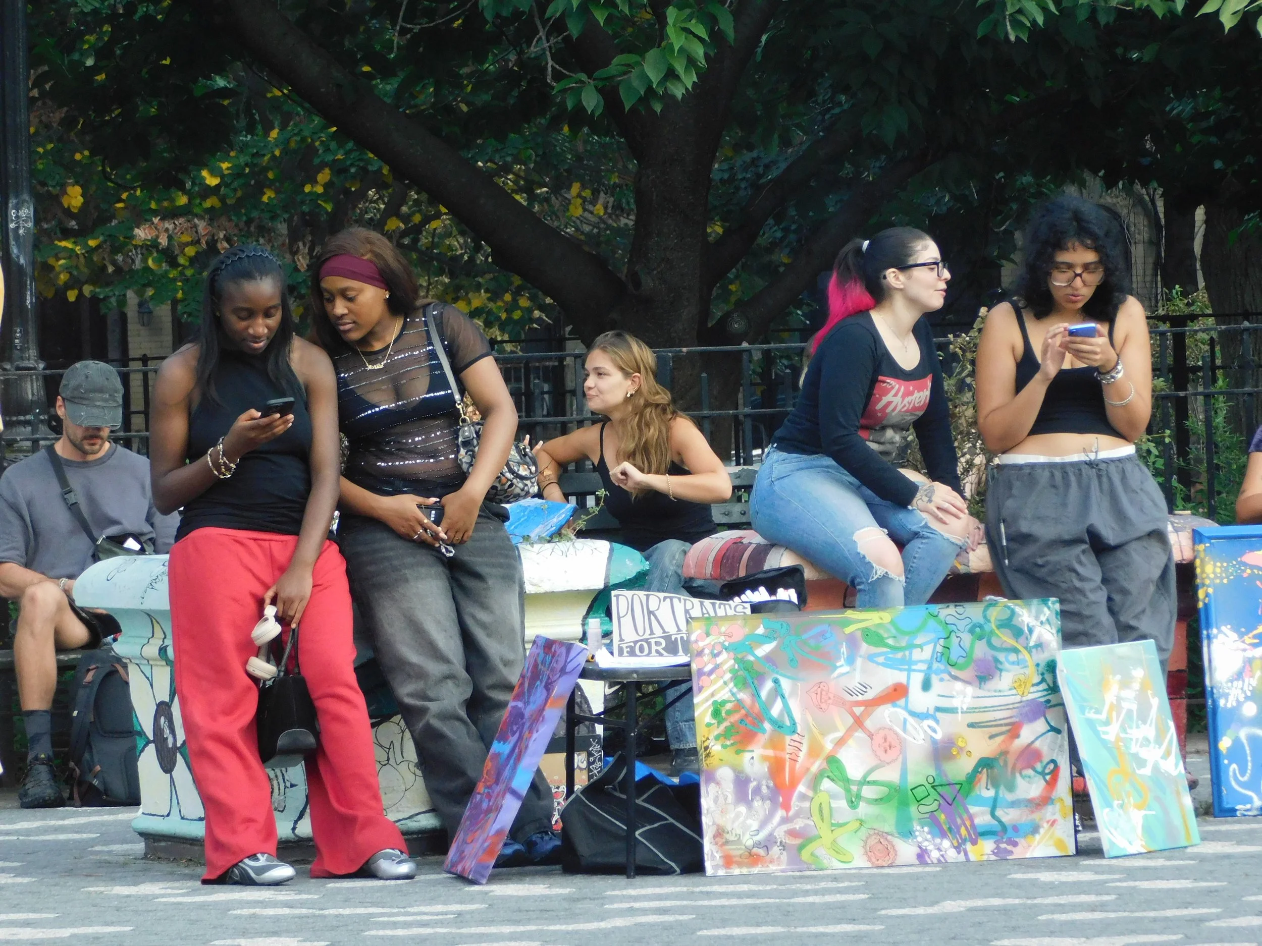 Group of five women sitting and standing in a park with trees in the background. Some are looking at their phones, and behind them are colorful paintings and signs, including one that reads 'PORTRAITS'.