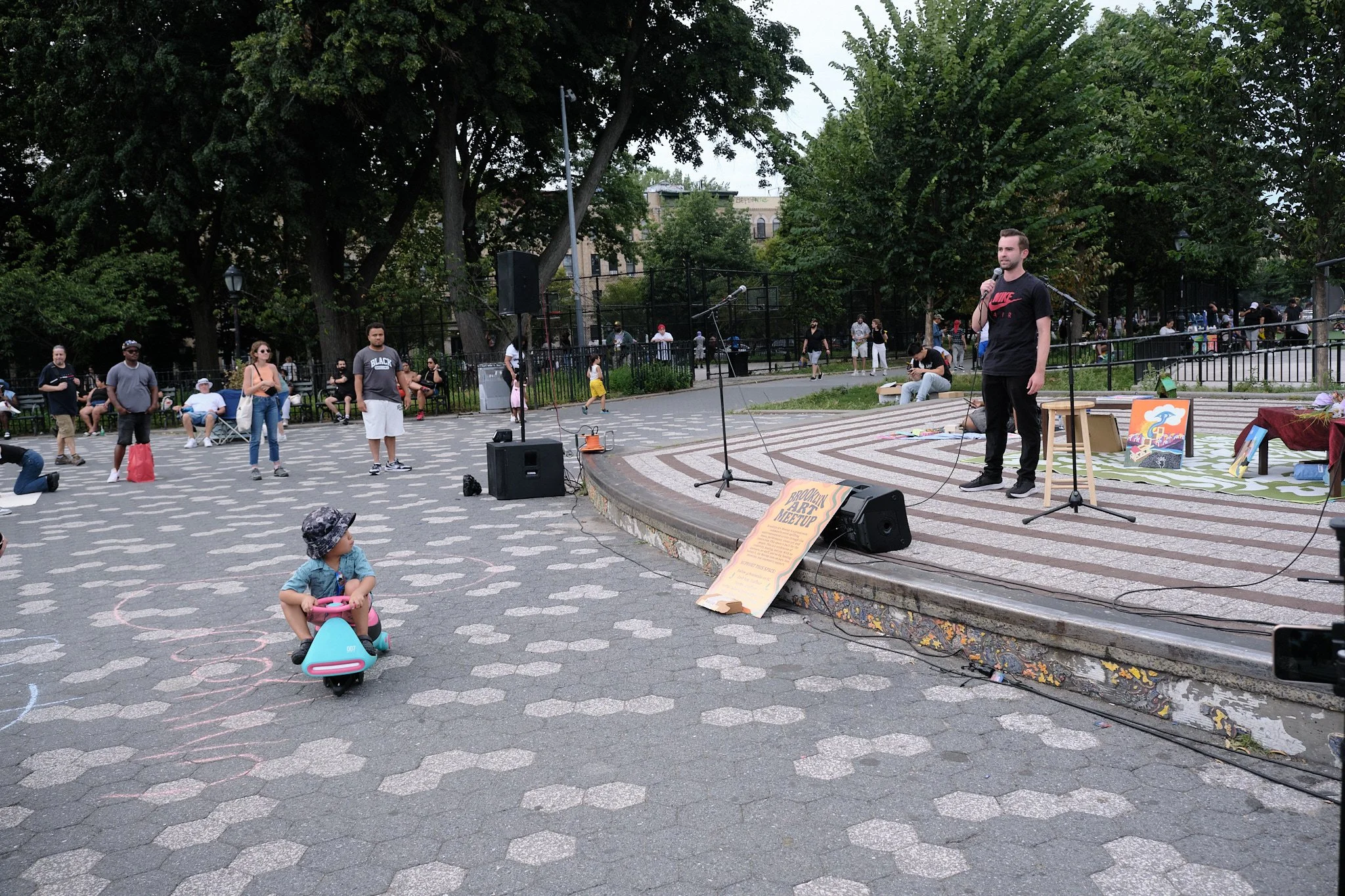 People gathered at an outdoor park for a community event or performance. A young man stands on a small stage with a microphone, while others sit or stand nearby. There are children playing, including a girl riding a pink and blue toy scooter. The bac