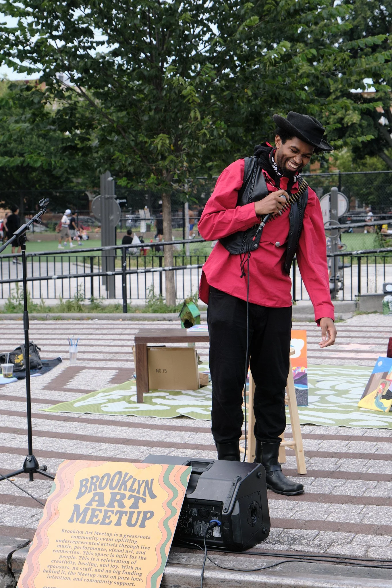 A man in a red jacket and black hat is smiling and holding a walkie-talkie. He is standing outdoors at a Brooklyn Art Meetup event, with a microphone and a large speaker nearby. There are trees, a park with people, and a fence in the background.