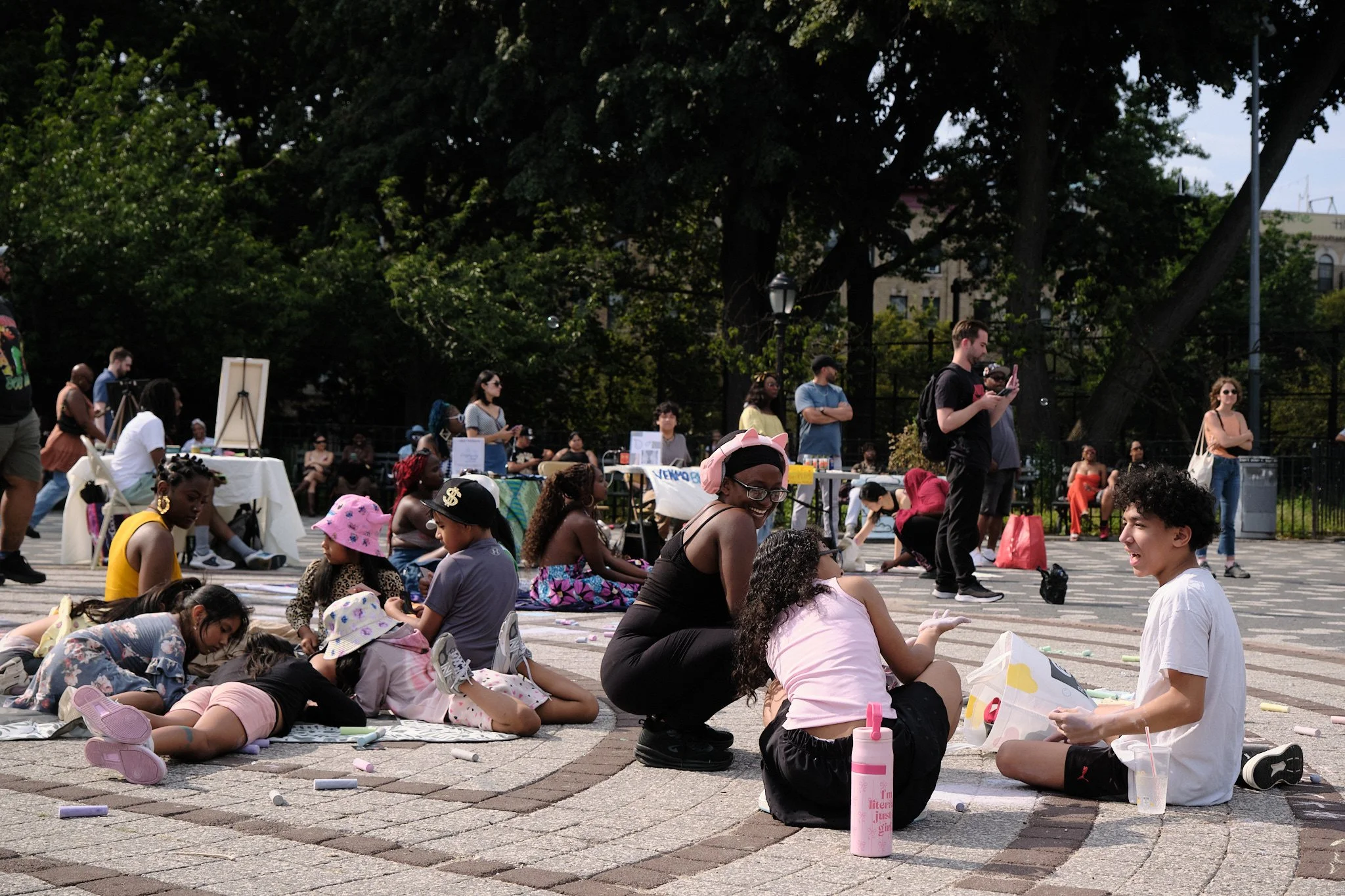 People gathered in a park, some sitting on the ground and others standing, with art supplies and signs around them, under large trees on a sunny day.