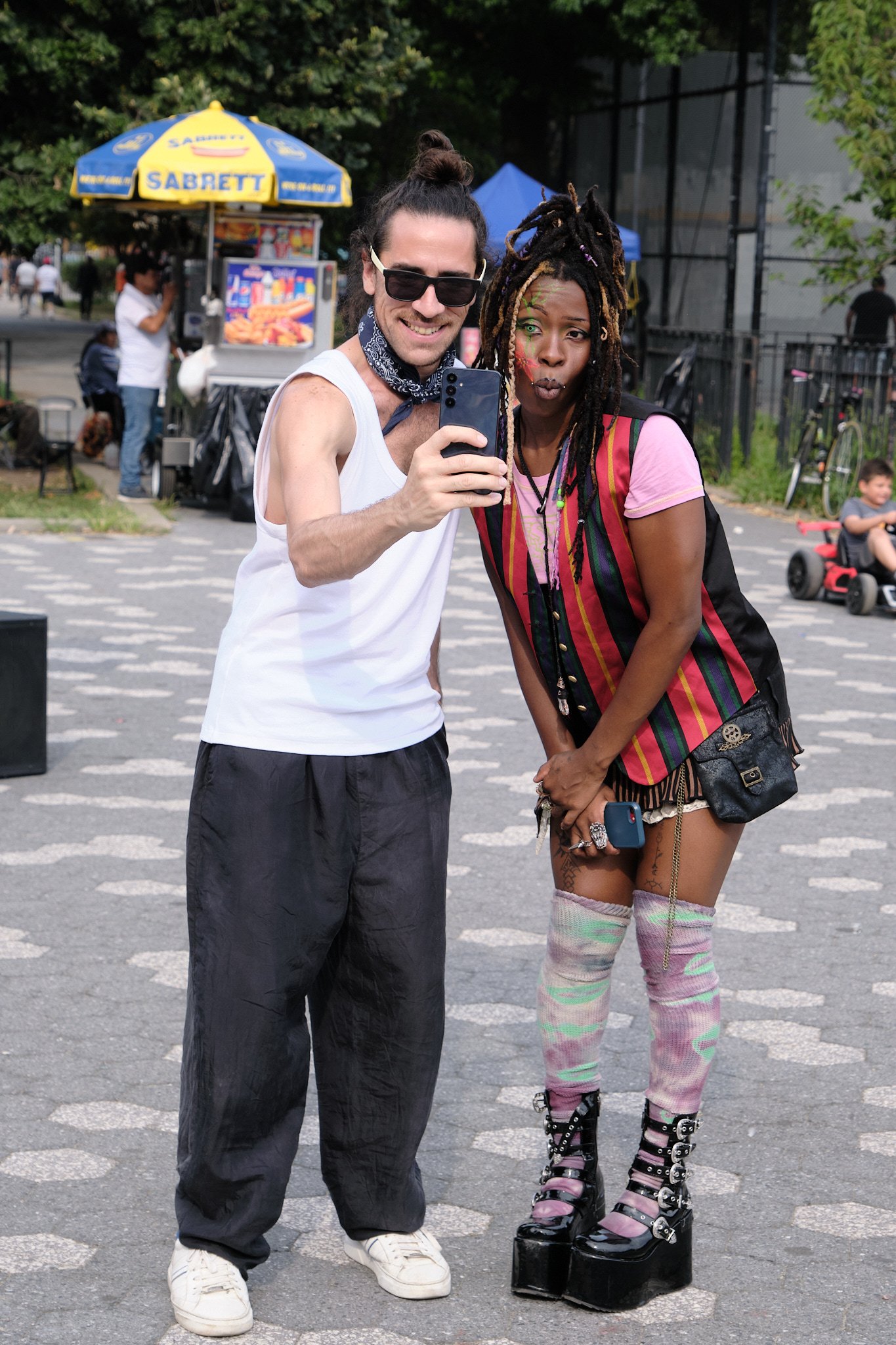 Two people taking a selfie in a park with trees, food cart, and other people in the background. One wears a white tank top and dark pants, and sunglasses. The other wears layered clothing, striped socks, platform shoes, and has colorful face paint and dreadlocks.