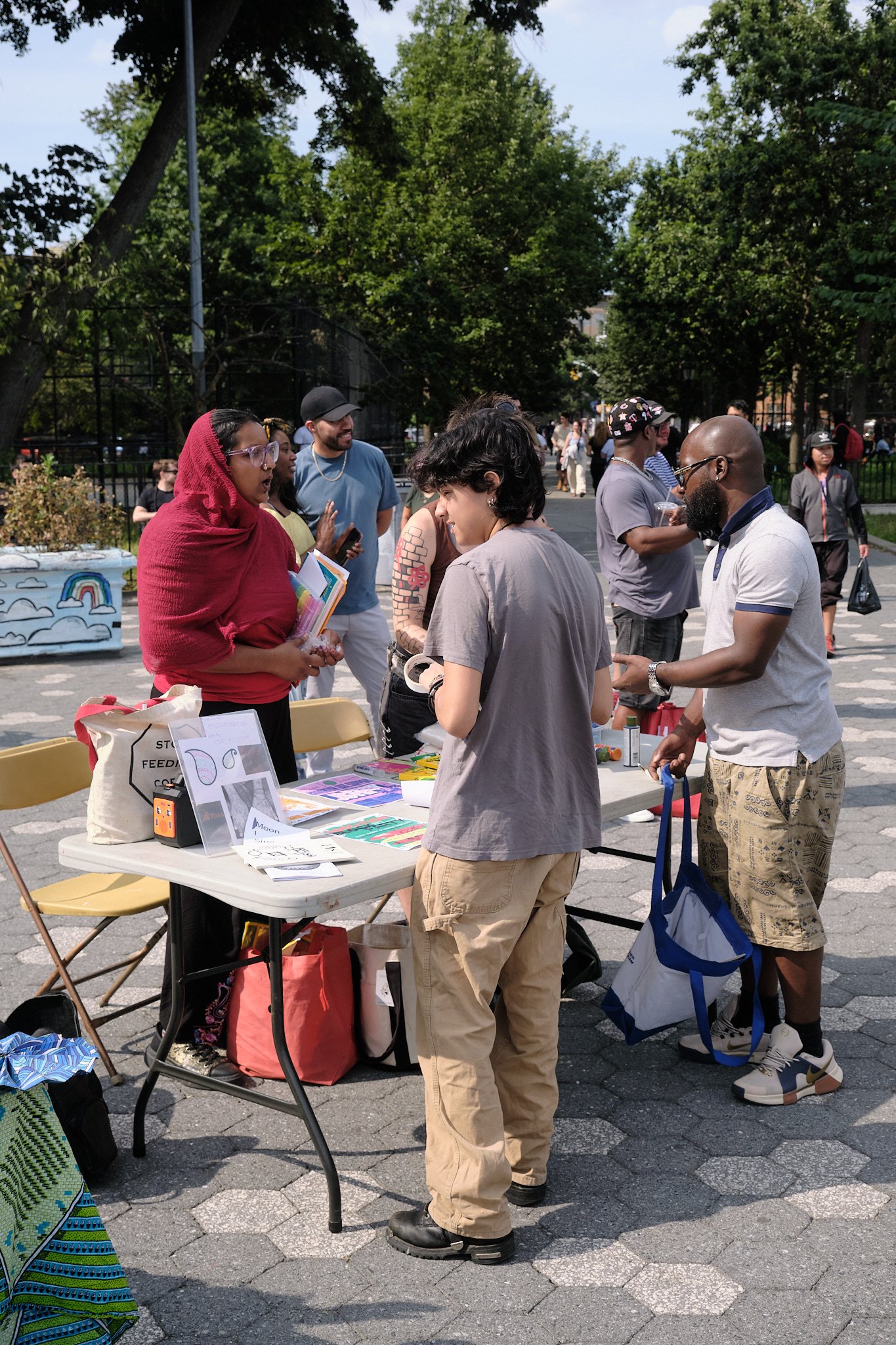People gather around a table outdoors in a park, engaging in conversation and browsing materials during a community event.