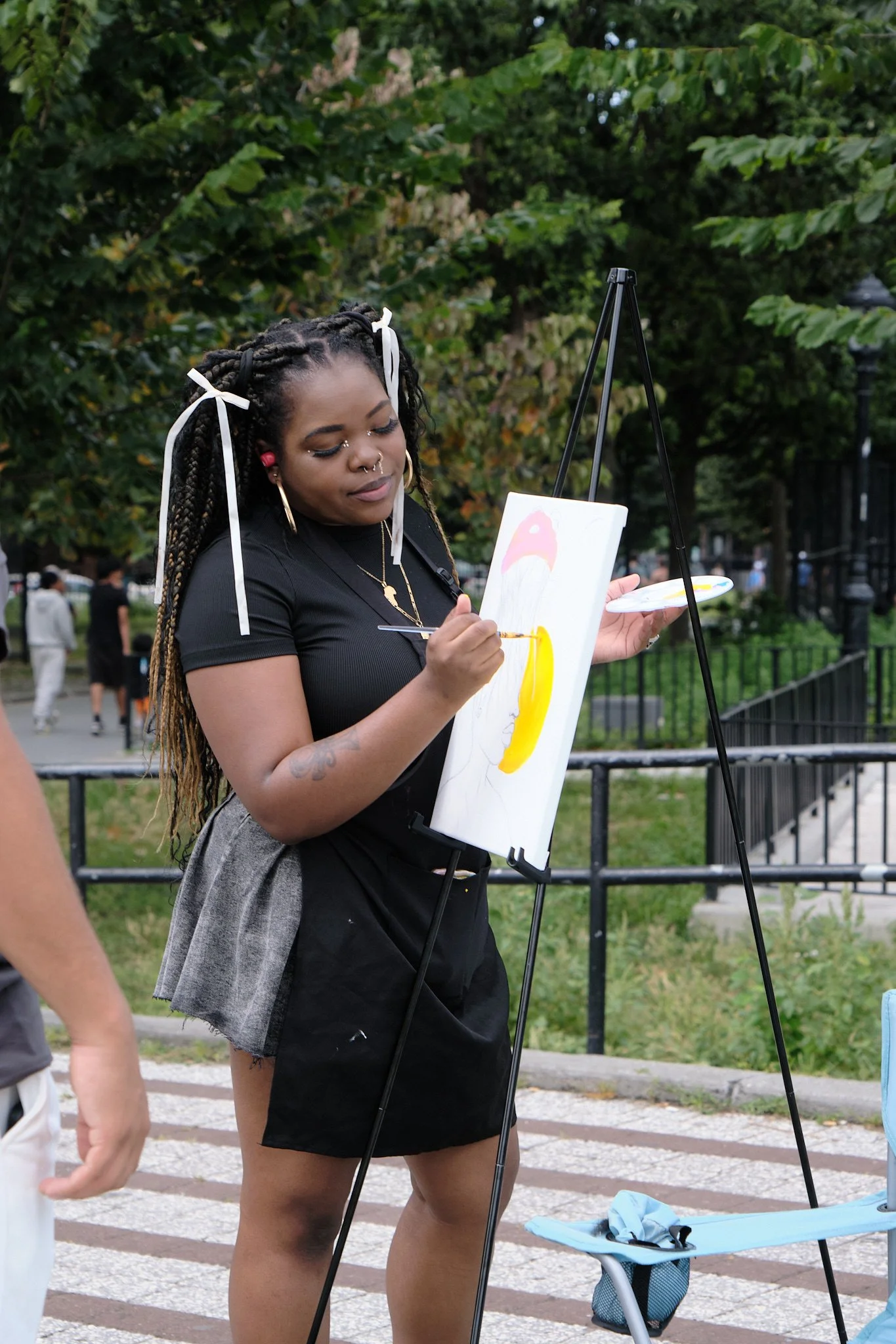 A woman with braids and face jewelry painting on a canvas outdoors in a park.