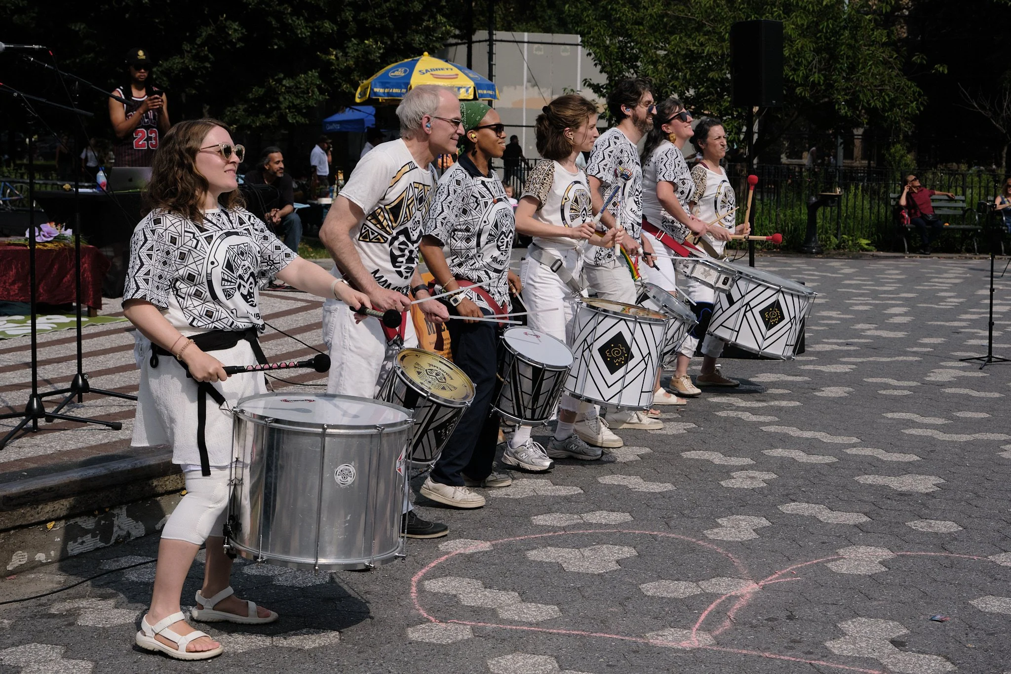 Group of people playing drums at an outdoor event in a park on a sunny day.