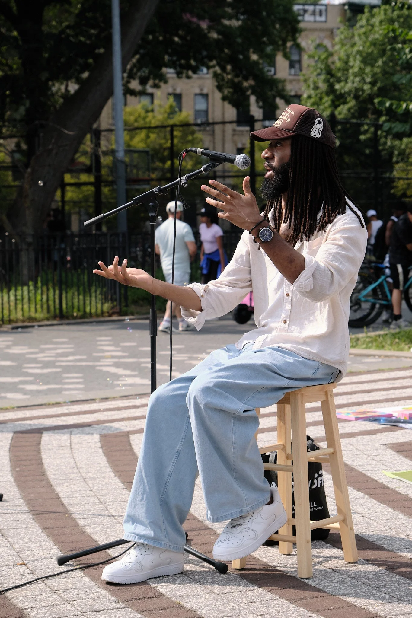 A man with long dreadlocks wearing a baseball cap, white shirt, and light blue jeans sits on a wooden stool in an outdoor setting. He is speaking into a microphone placed on a stand, with several people in the background and trees and a building behi