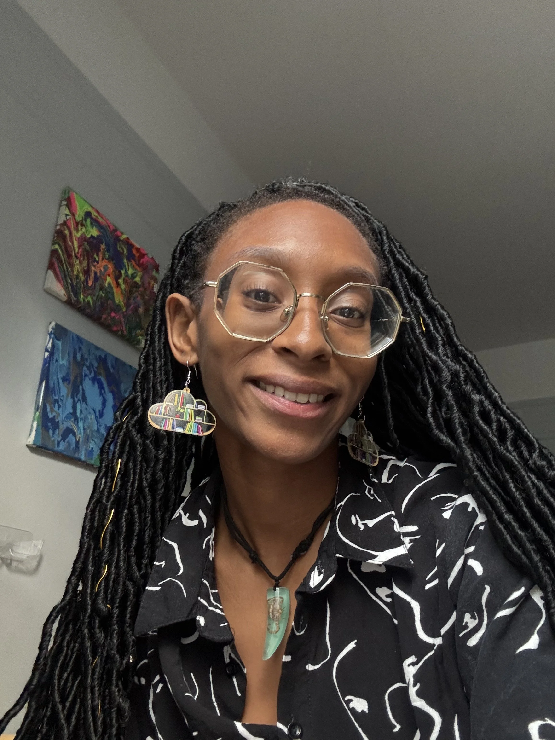 Close-up of a woman with long braided hair, wearing glasses with geometric frames, colorful cloud-shaped earrings, a jade pendant necklace, and a black and white patterned blouse, smiling at the camera. Background shows abstract paintings on a wall.