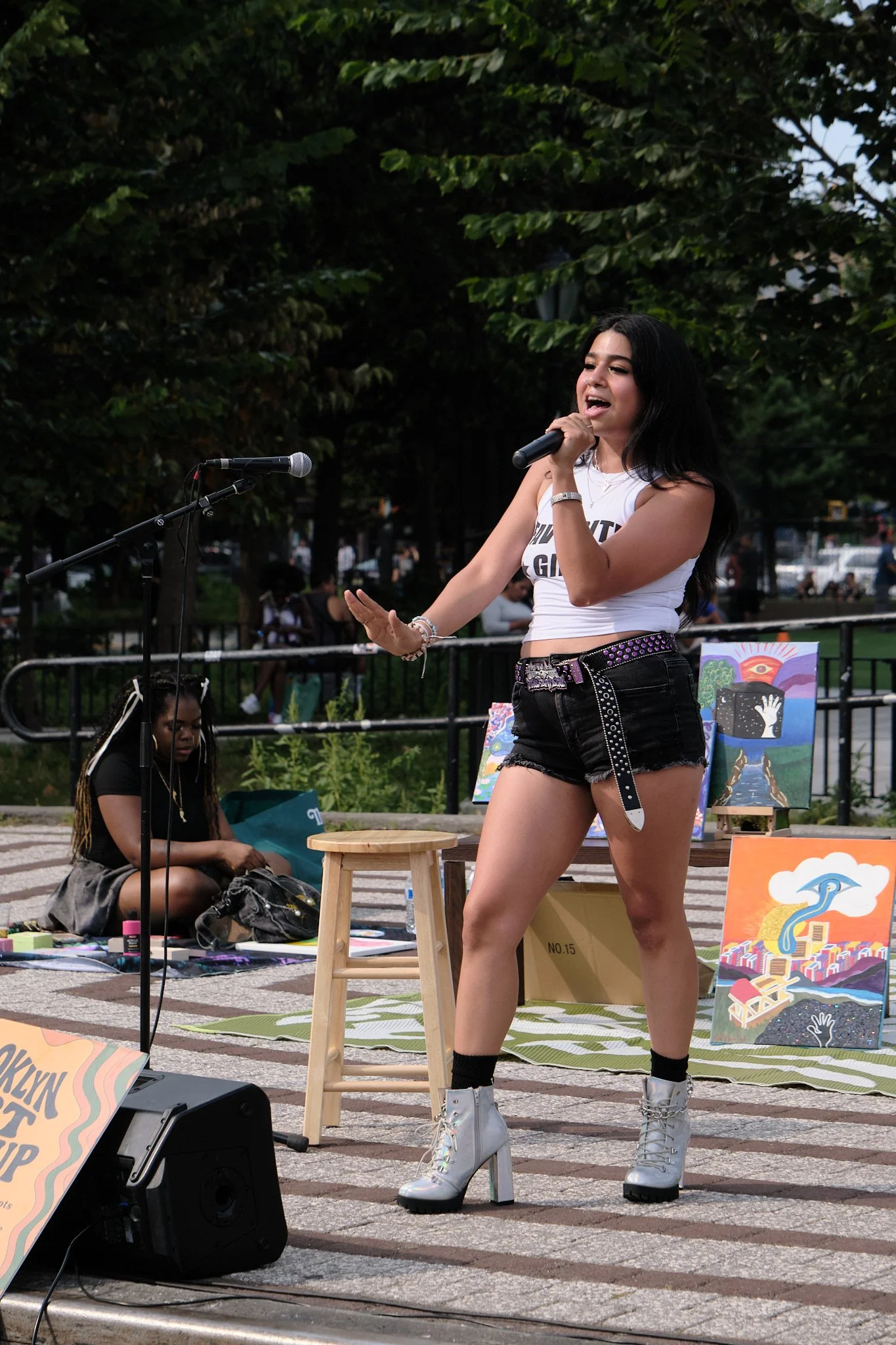 A woman with long dark hair singing into a microphone at an outdoor event, wearing a white sleeveless top, black shorts with silver studded belt, white lace-up boots, and black socks. Another woman with braided hair and glasses is sitting on the grou
