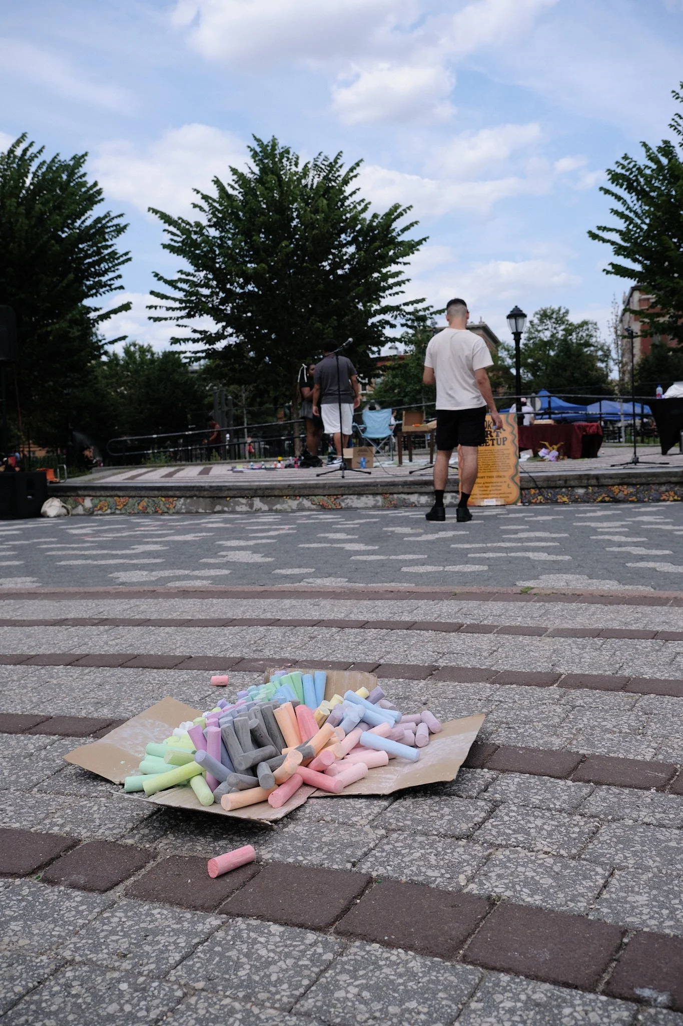 A pile of pastel chalks on a piece of paper in the foreground, with an outdoor event stage and people preparing in the background on a cloudy day.