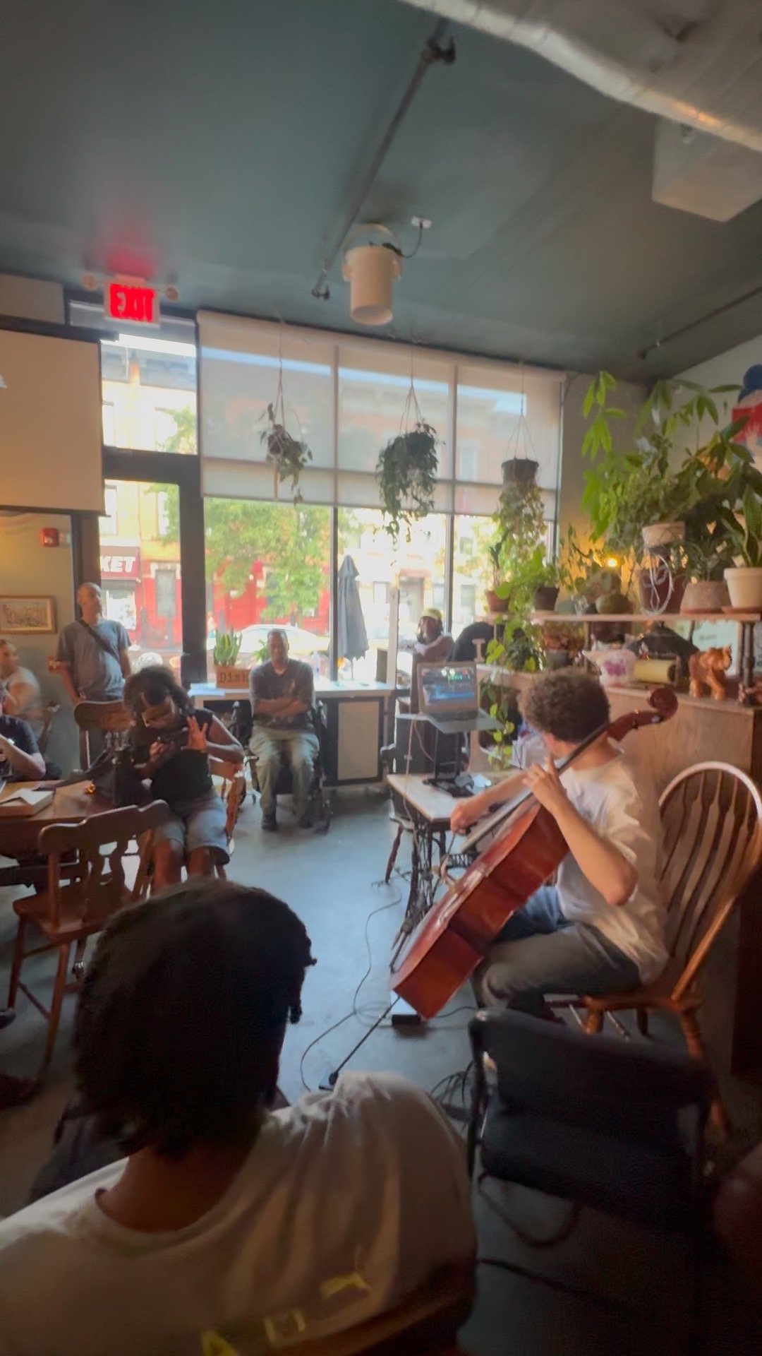 A group of people gathered inside a cozy cafe watching a live musical performance featuring a cellist and a guitarist.
