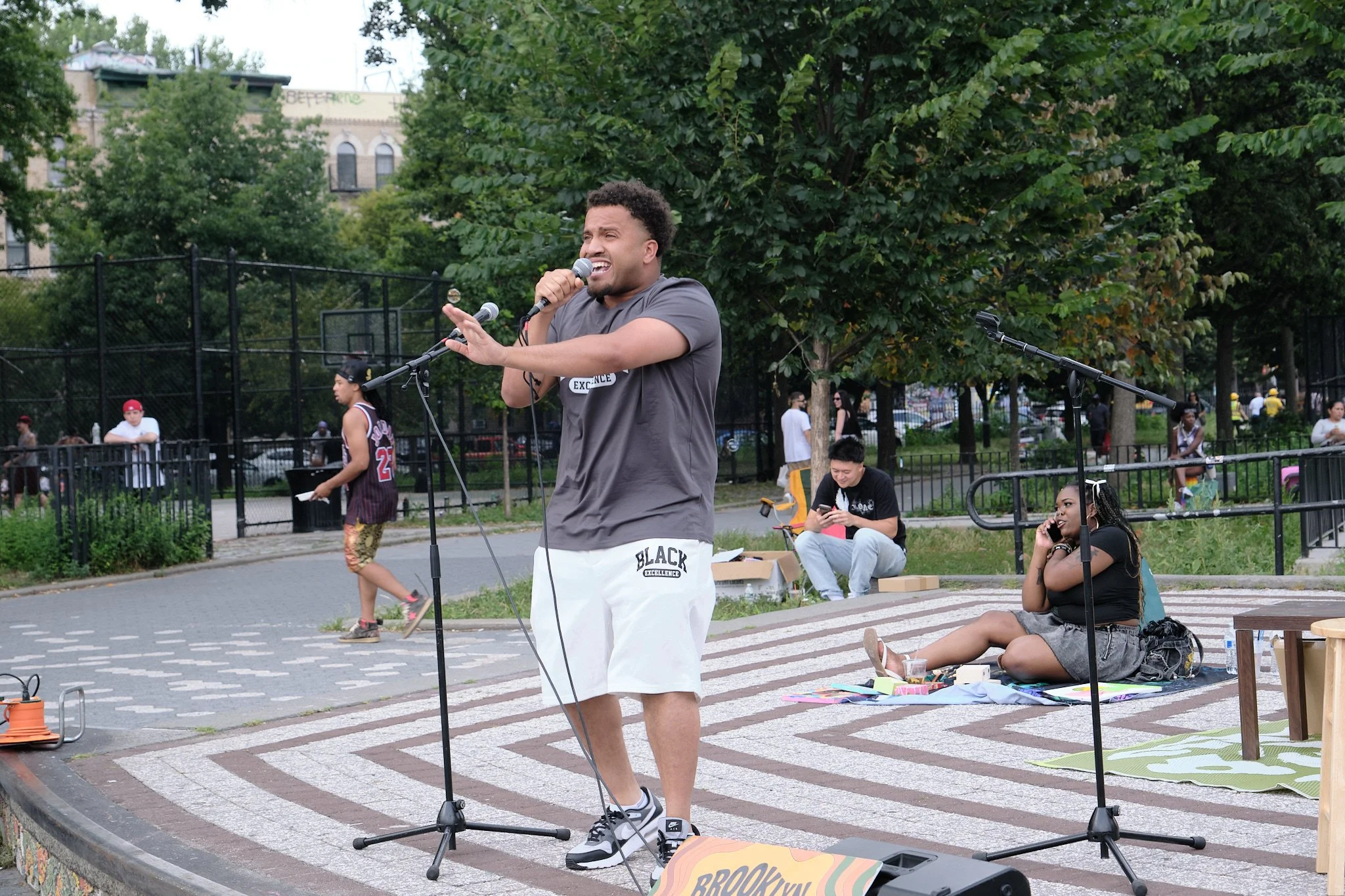 A man singing into a microphone outdoors in a park, with people sitting and walking around, and a basketball court in the background.