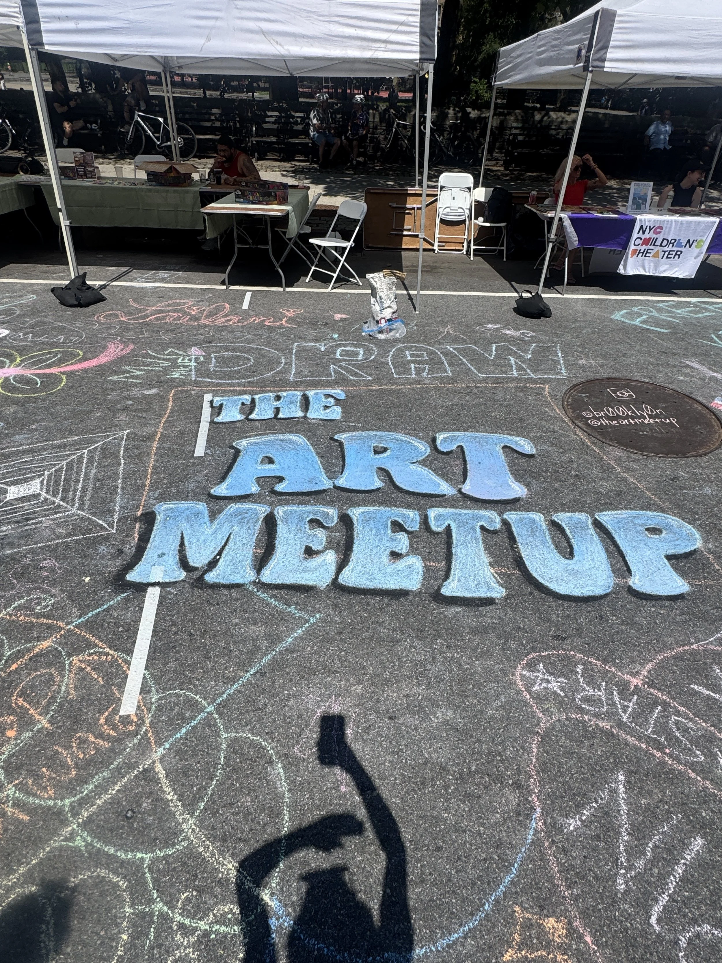 Street scene with tents and tables at an outdoor event, featuring colorful sidewalk chalk art and a manhole cover, with people sitting at booths in the background.