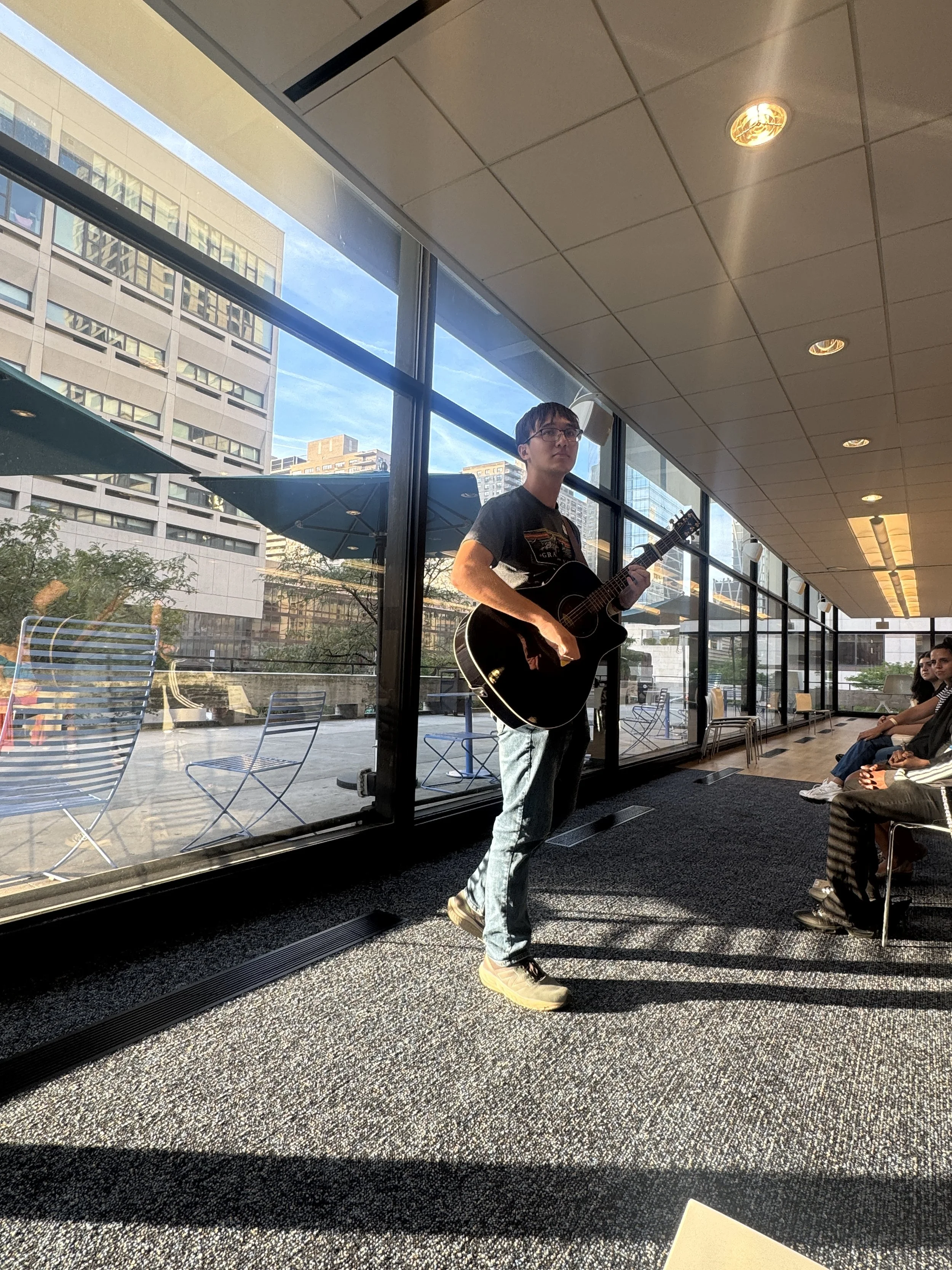 A man playing an acoustic guitar while standing indoors near large glass windows showing a cityscape with tall buildings and outdoor seating. Several seated people are visible, with sunlight streaming in.