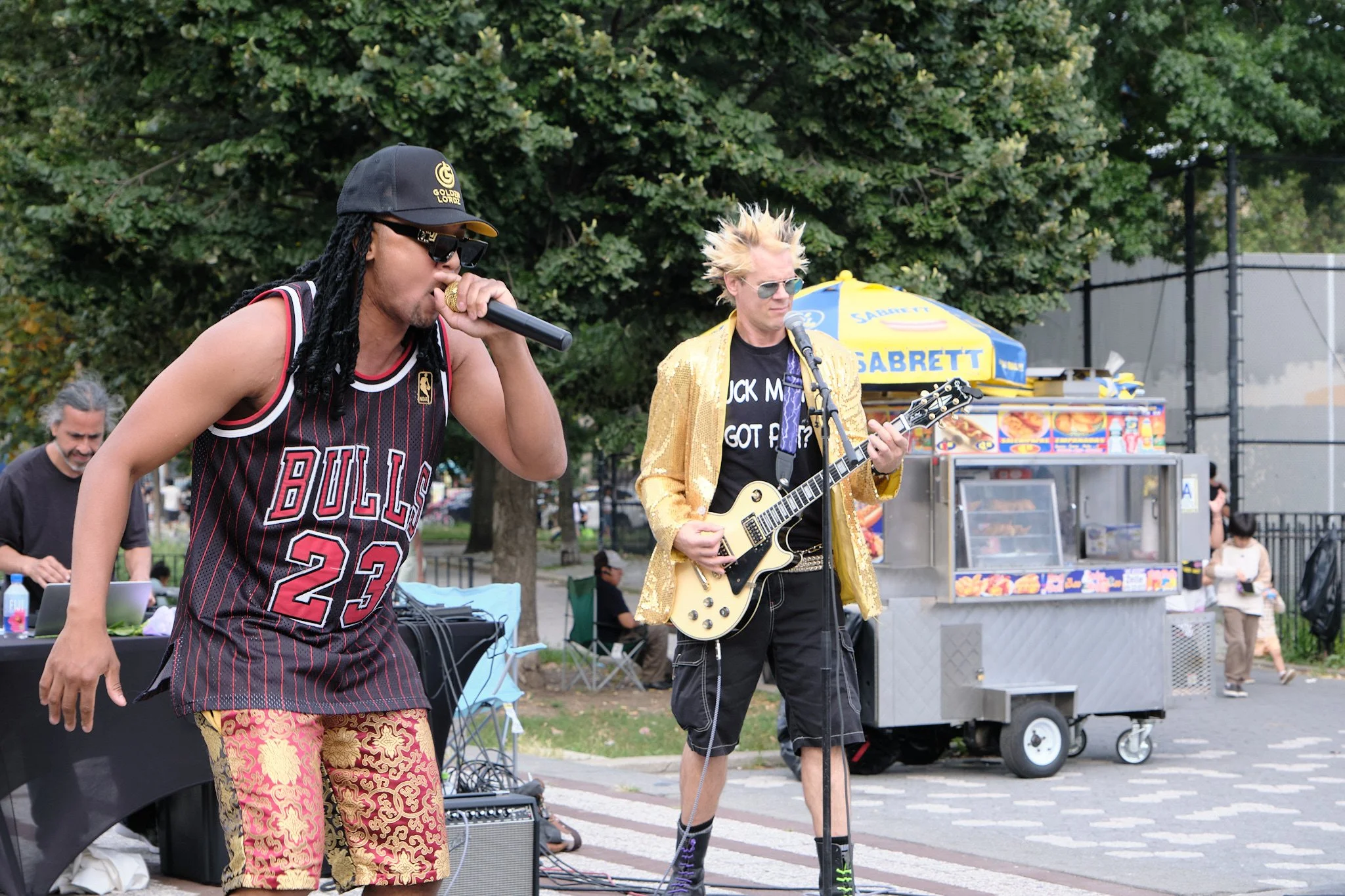 Two male musicians performing outdoors in a park, one singing into a microphone and wearing a Chicago Bulls jersey, the other playing an electric guitar and dressed in a bright gold jacket and sunglasses, with a food stand and trees in the background