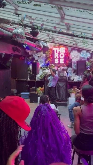 A performer on stage at a nightclub or dance club with a DJ setup, smoking, and a large screen that says 'THE ROSE'. Audience members are facing the stage, with one person in the foreground wearing a red hat and another with bright purple hair.