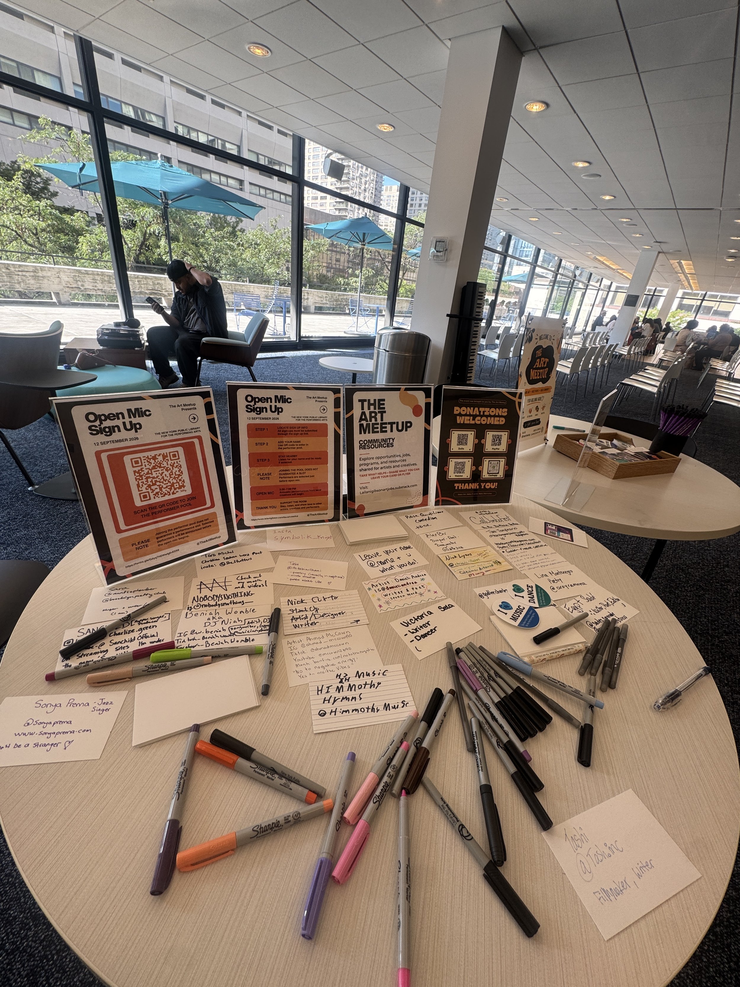 A round table with various pens, markers, and handwritten notes, with informational signs about an art meetup and open mic sign-up in the background. The room has large glass windows showing an outdoor patio with tables, umbrellas, and a cityscape in the distance.