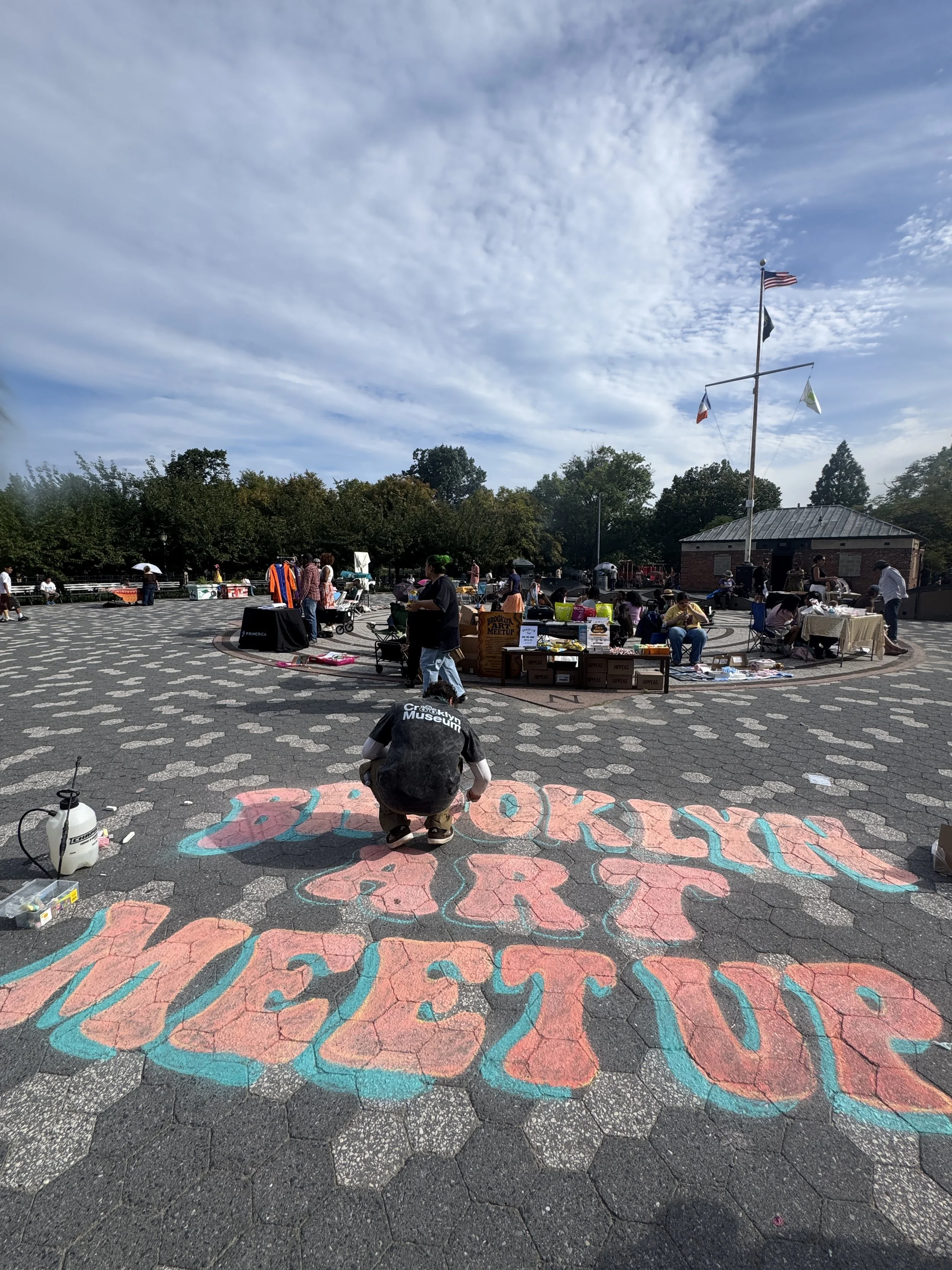 People gathered at an outdoor event with vendors and children, under a partly cloudy sky, with an American flag flying on a tall pole in the background. A person is creating colorful chalk art on the ground that says 'Brooklyn Museum'.