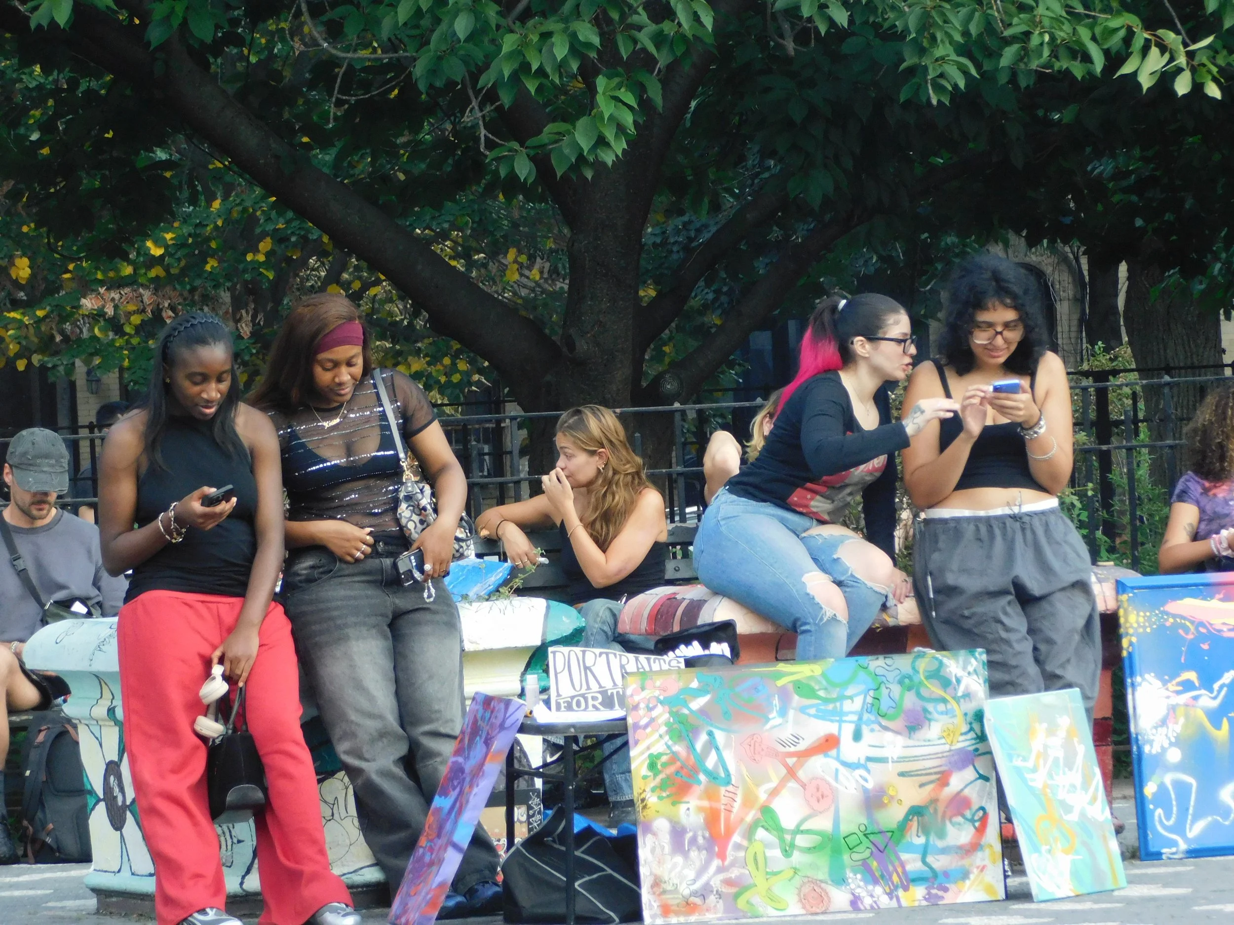 Group of diverse women at an outdoor event, some looking at their phones, surrounded by colorful paintings and trees.