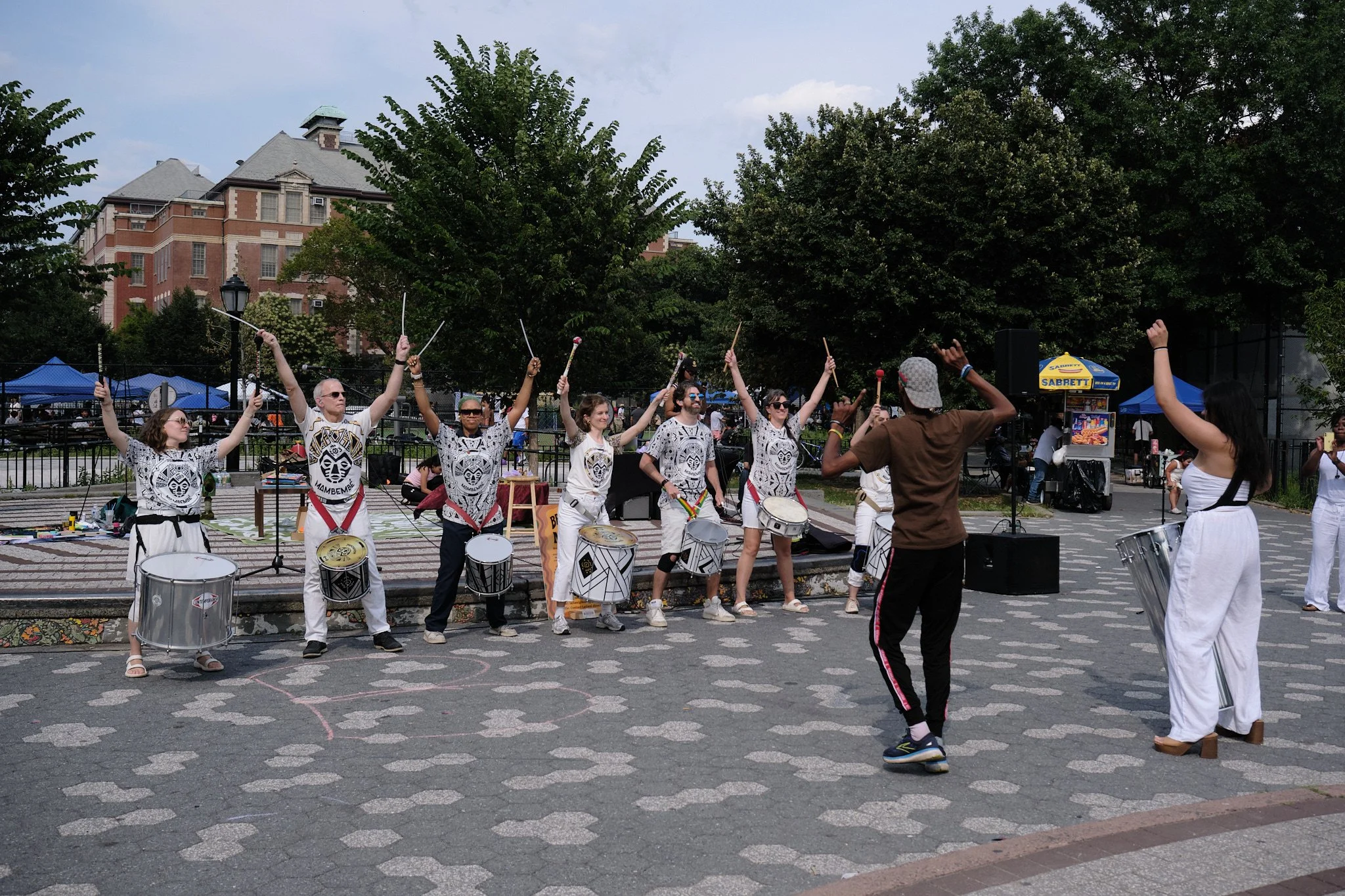 Group of musicians playing drums and percussion instruments in an outdoor park setting with trees and a historic brick building in the background, during a sunny day.
