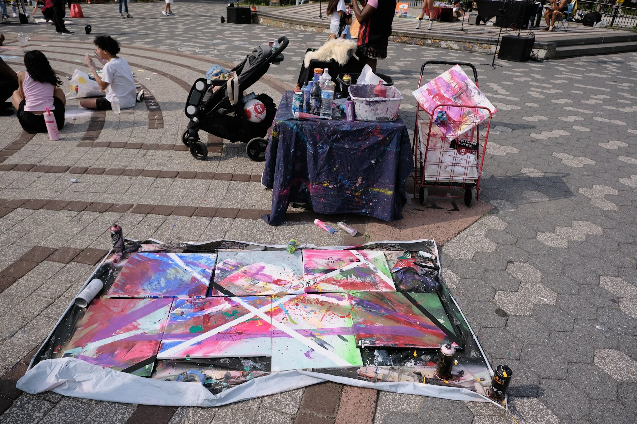 Outdoor scene of an art vendor's station at a public event, featuring painted canvases, paint bottles, spray paint, and a cart with art supplies on a paved area. In the background, people are sitting and standing near a stage or platform, and childre