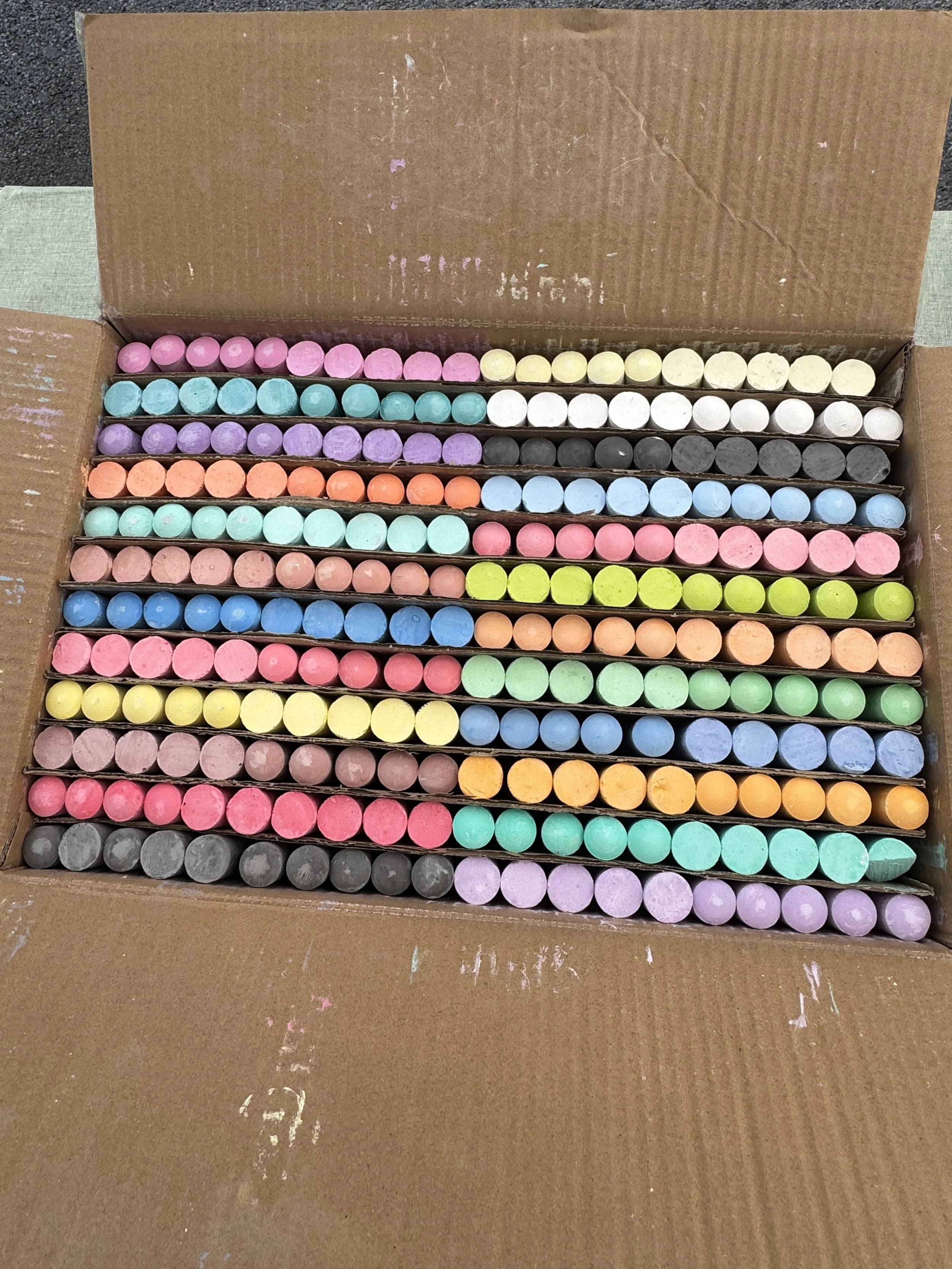 A cardboard box filled with rows of colorful chalks in pastel shades of pink, blue, purple, black, white, orange, yellow, and green.