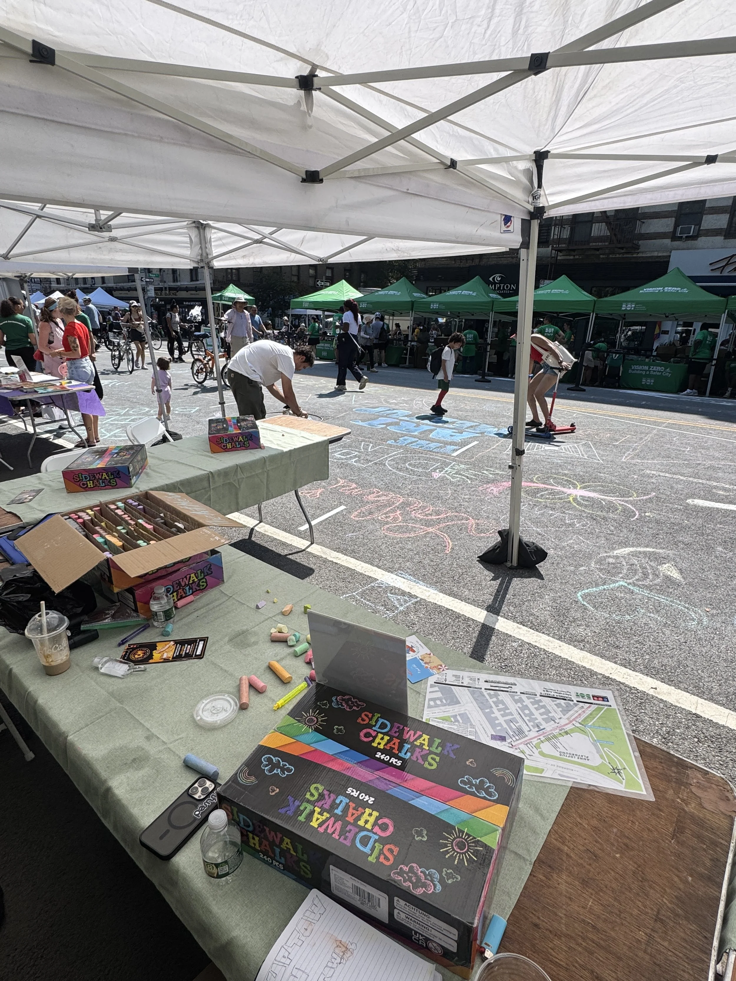 Street fair with tents, chalk art on pavement, people walking, children playing, tables with sidewalk chalk boxes, flyers, and maps.