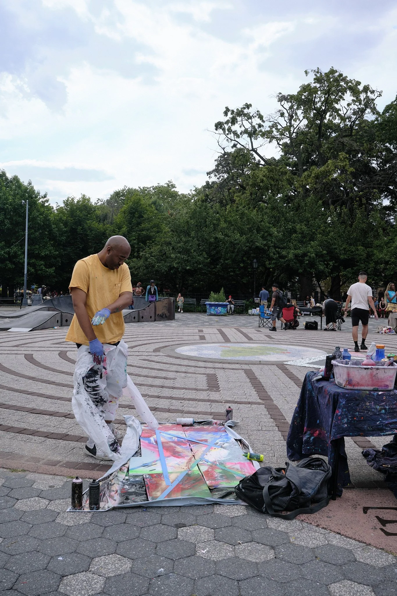 A man in a yellow t-shirt and white paint-splattered pants is painting in a city park with multiple people in the background. He is standing on a painted canvas on the ground. Nearby are cans of spray paint and other art supplies, and artist's equipm