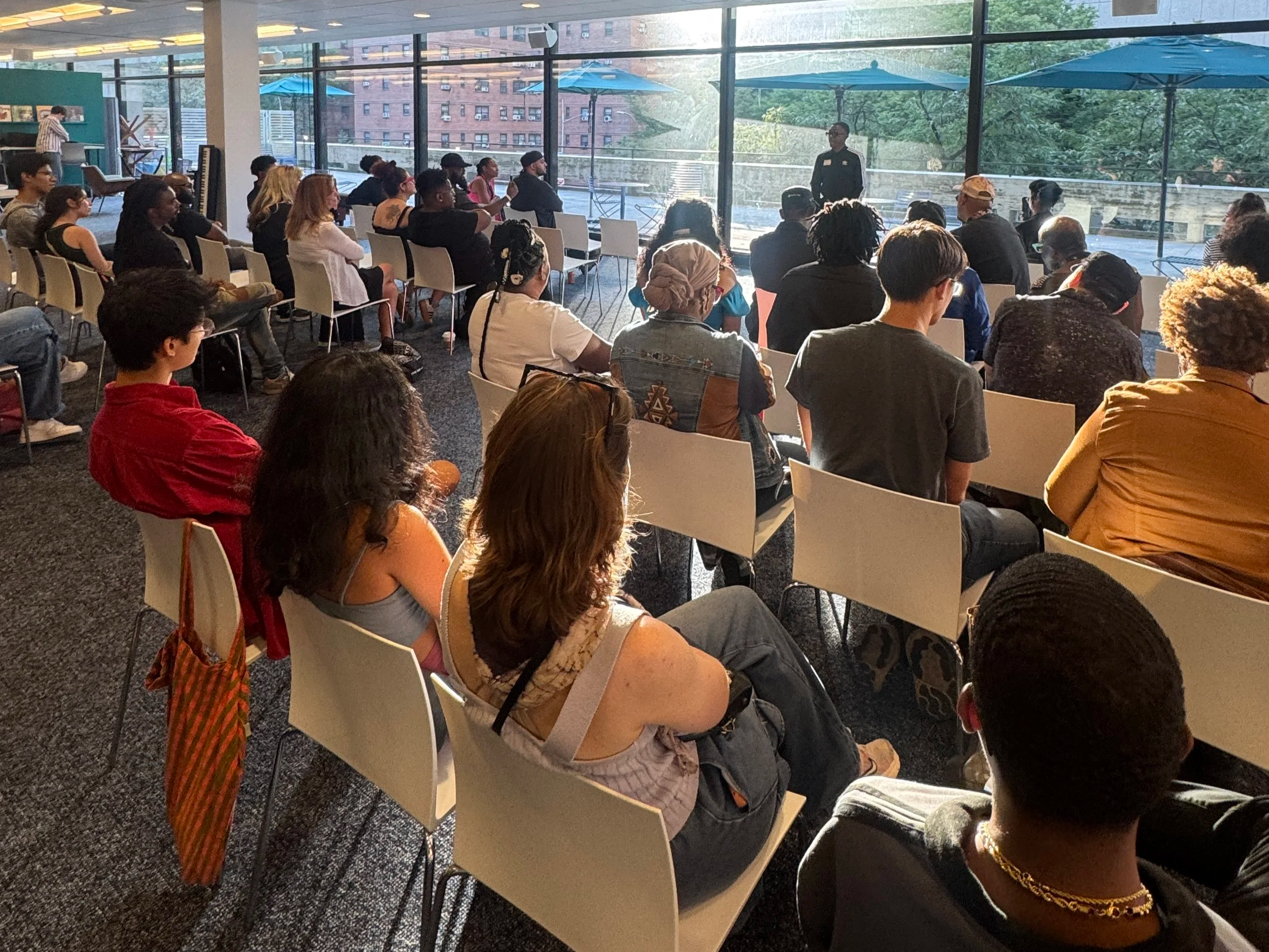 A group of diverse people sits in a room with large floor-to-ceiling windows, attentively listening to a speaker standing at the front. The room has a bright, natural light and views of buildings and trees outside.