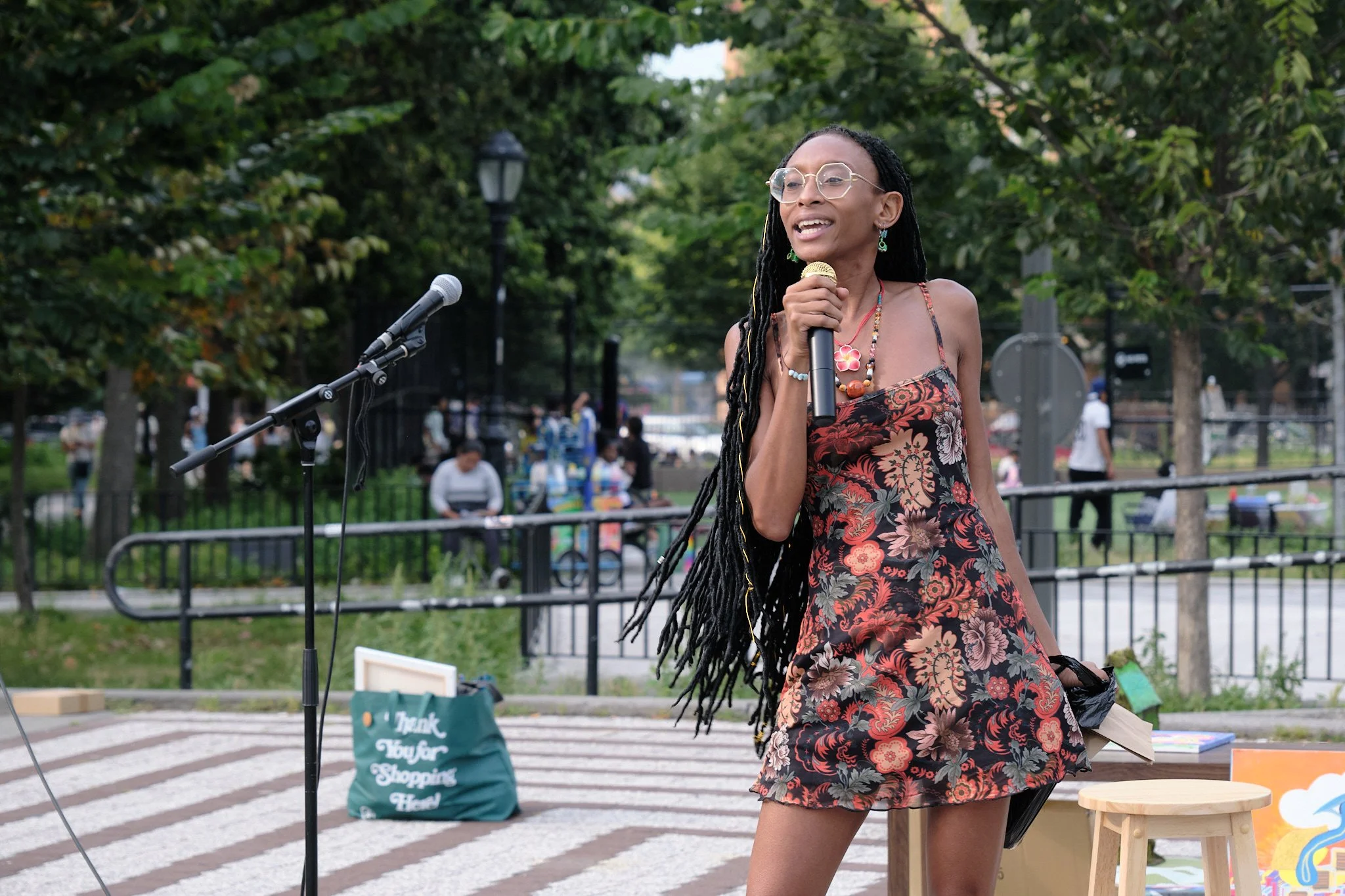 A woman with long dreadlocks, wearing glasses, colorful earrings, and a floral dress, speaking into a microphone outdoors in a park with trees and people in the background.