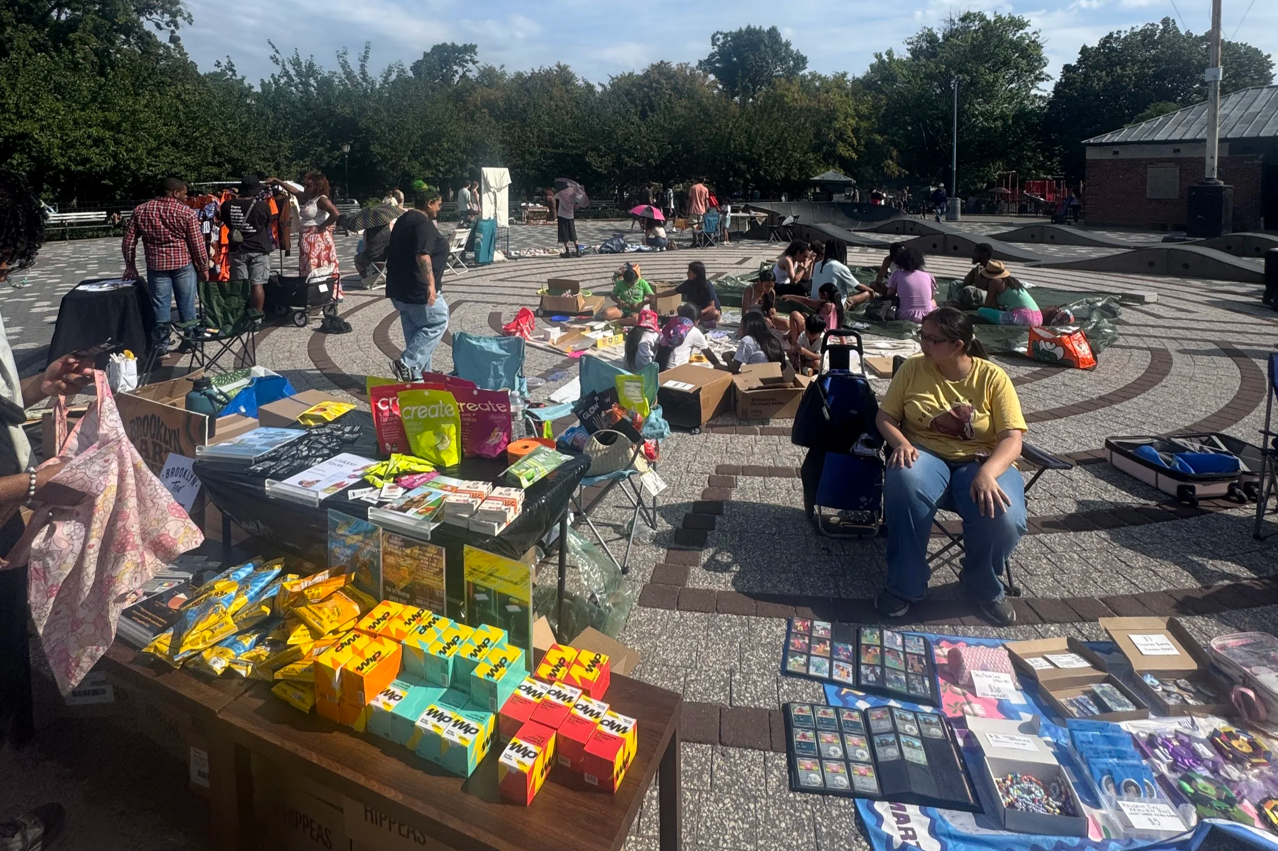 A busy outdoor flea market or street fair with tables displaying various items like books, toys, and collectibles. People browse, chat, and explore the booths, some sitting on the ground or in chairs. Trees and a small brick building are visible in the background.