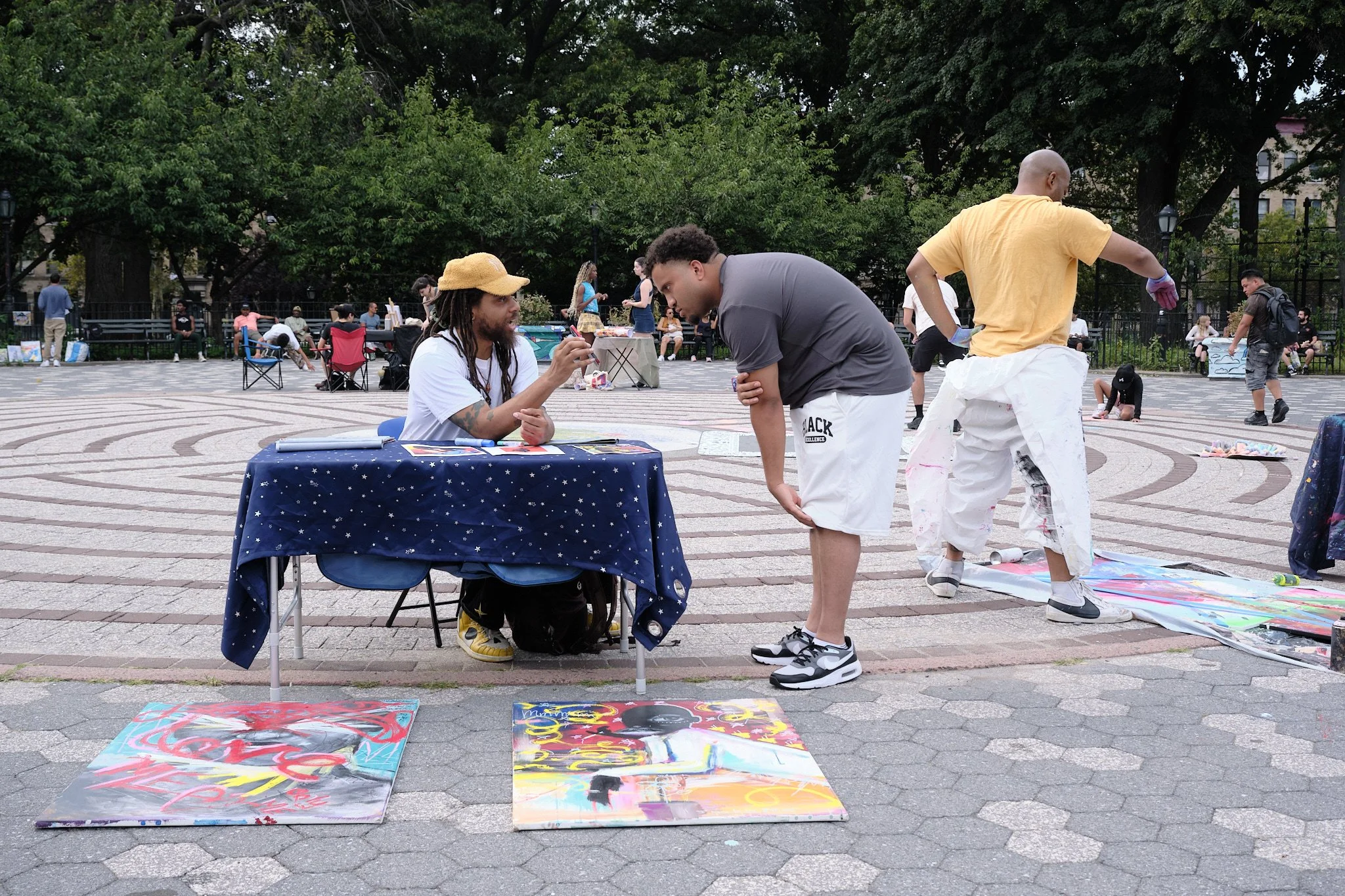 People at an outdoor art fair with paintings on display on the ground, two men talking at a table, and others in the background, surrounded by lush green trees.